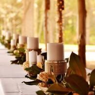 Row of white candles with greenery and woven holders on a long table by a sunlit window