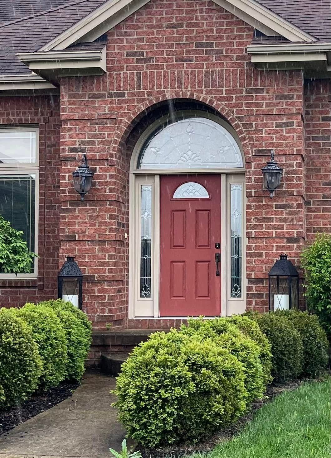 The front door of a brick house with a red door surrounded by bushes and lanterns.