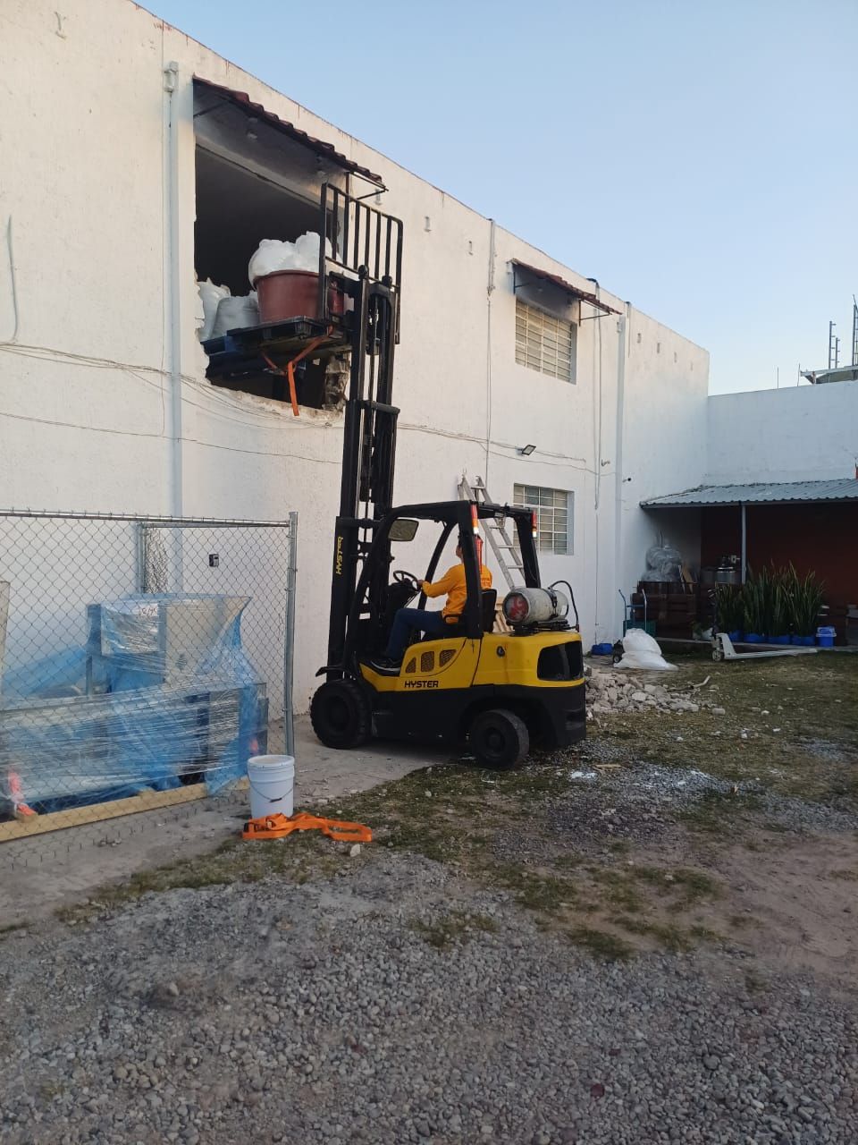Una carretilla elevadora amarilla levanta un palé hacia la ventana de un almacén, con palés y equipos en el exterior.