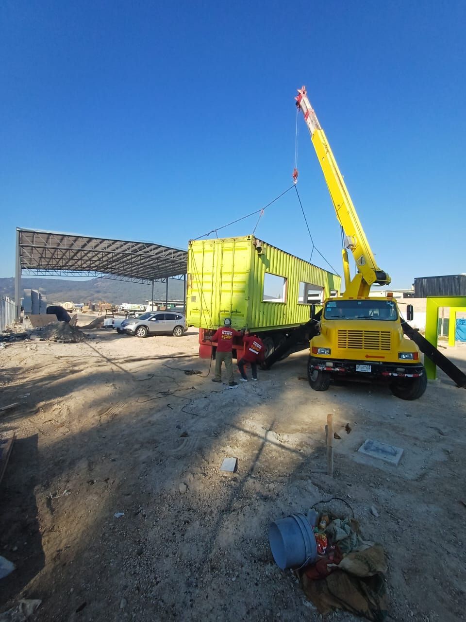 Una grúa amarilla levanta una cabina móvil de color amarillo brillante en una obra nevada bajo un cielo azul despejado.