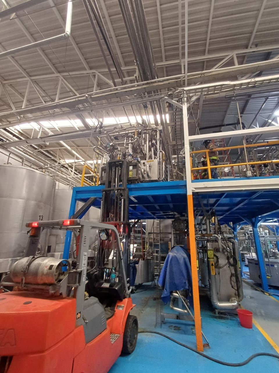 Carretilla elevadora en un almacén industrial bajo un altillo metálico, con maquinaria azul y postes de soporte naranjas.