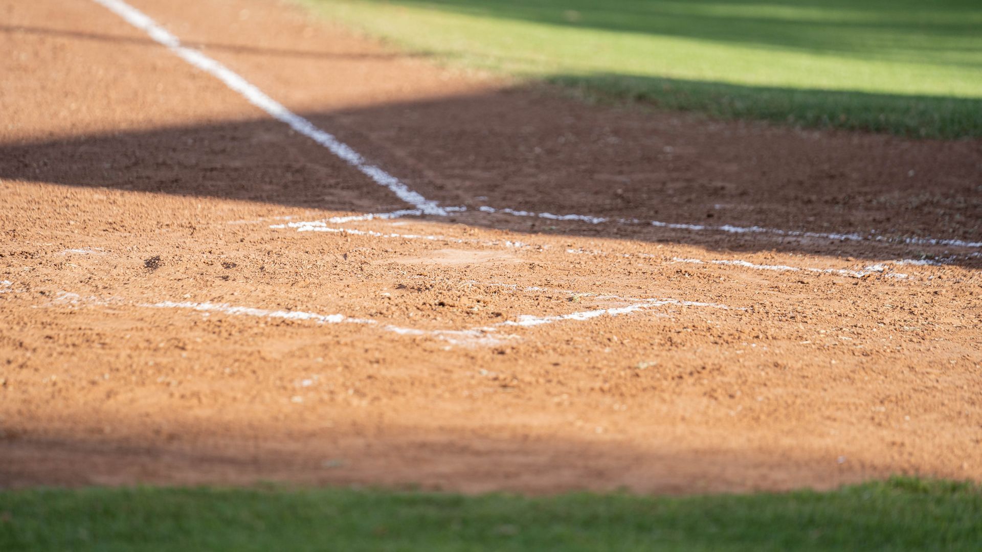 Baseball batter's box, infield dirt, chalk lines, and green grass in sunlight.