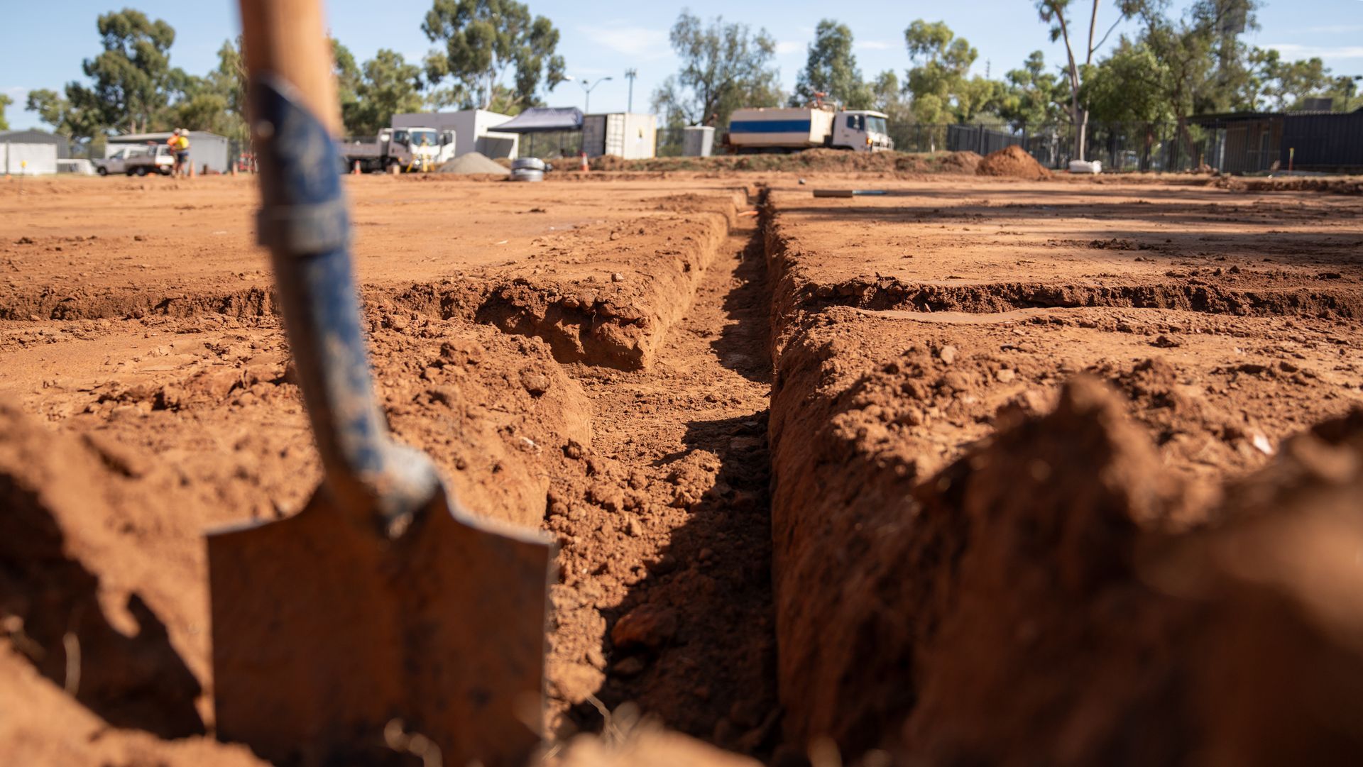 A shovel in a dirt trench at a construction site. Reddish-brown soil. Blue sky and buildings in the distance.