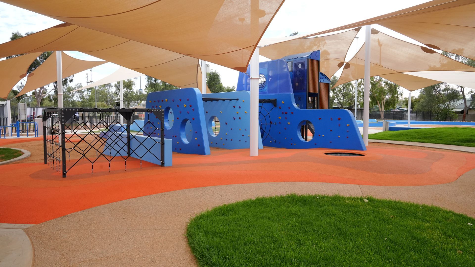 Children's playground with blue climbing structure, orange ground, tan shade sails, and green grass.