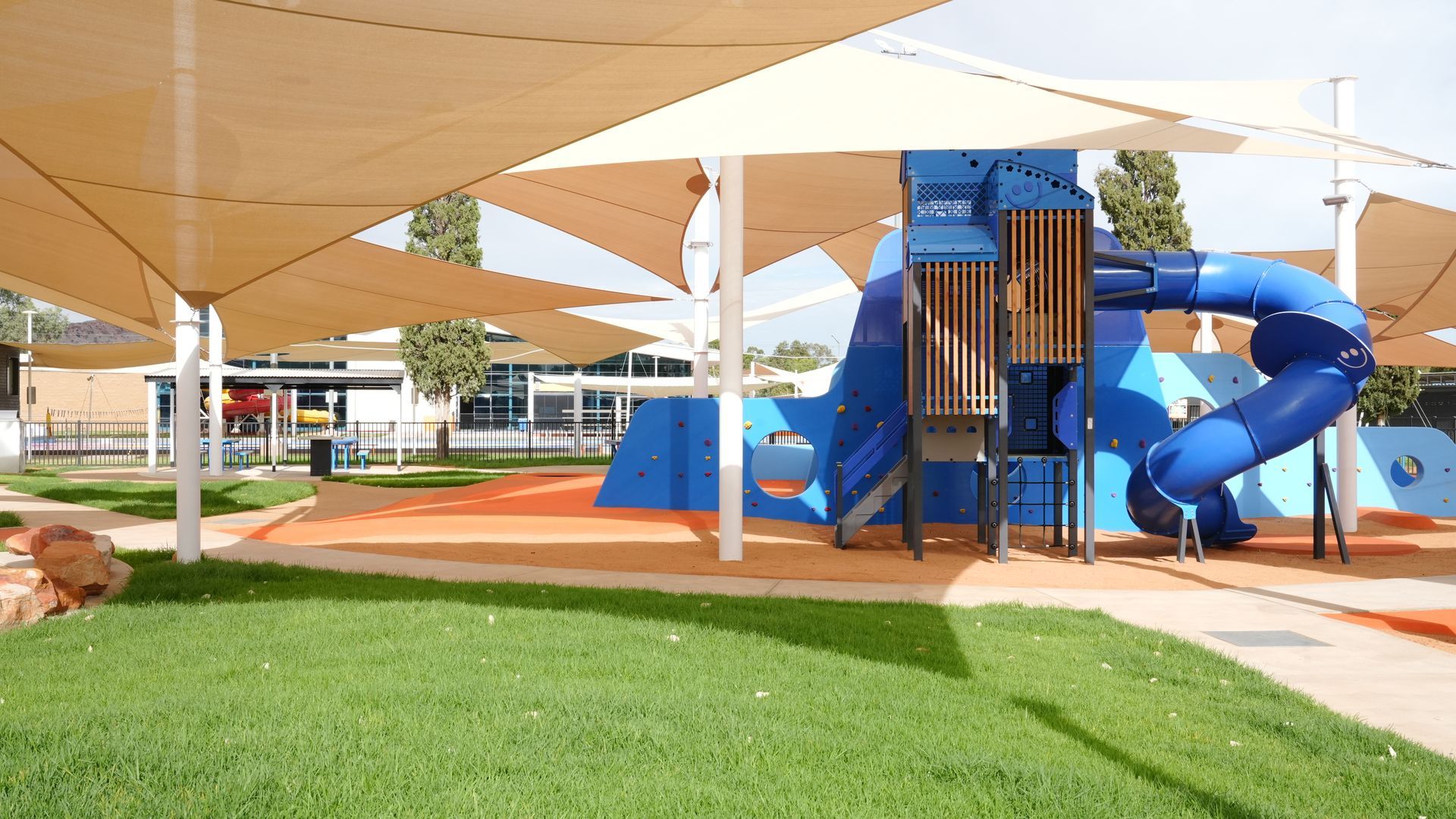 Playground with blue climbing structure, beige shade sails, and green grass.