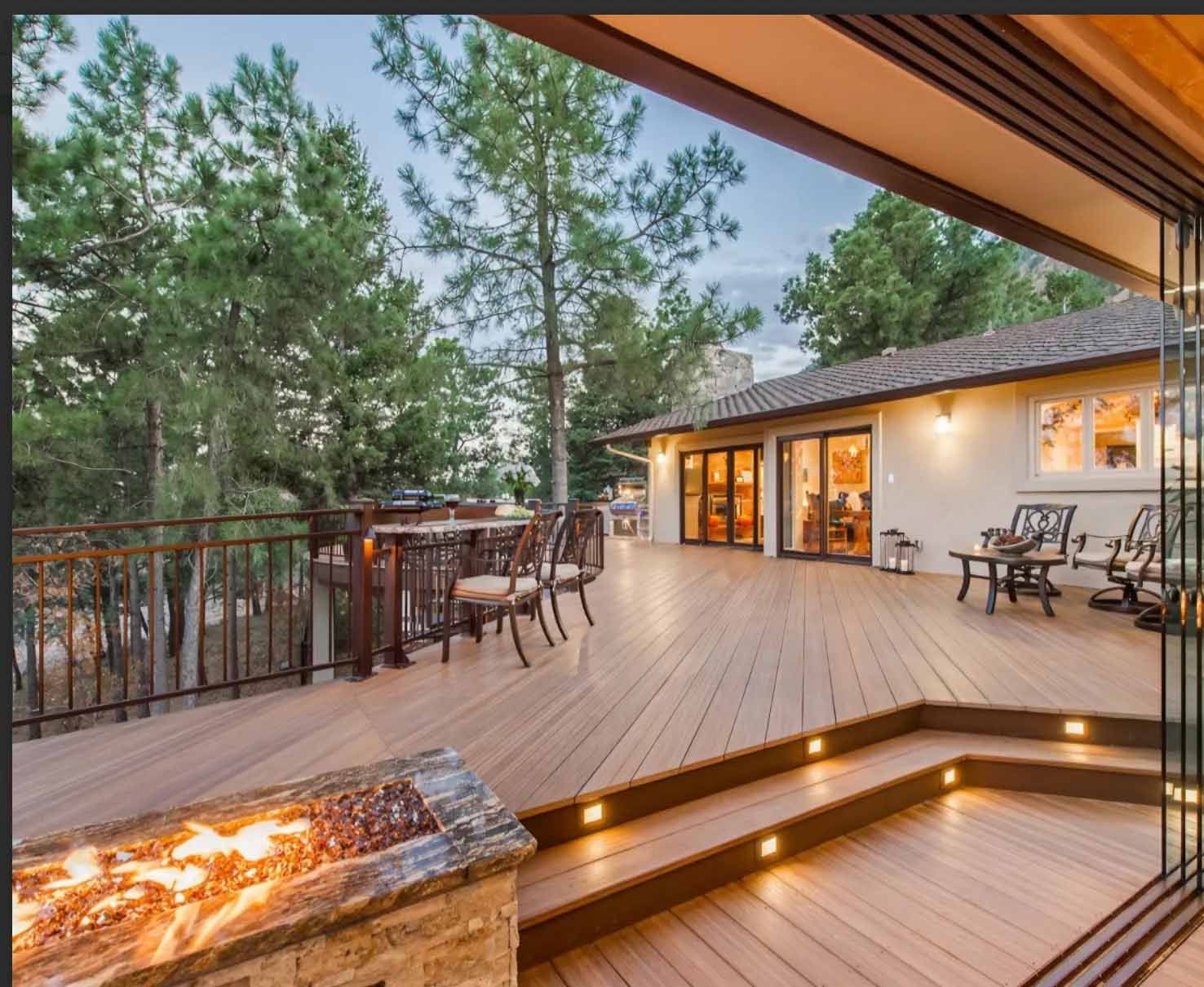 Wooden deck with fire pit and outdoor dining overlooking a home with glass doors, surrounded by trees.