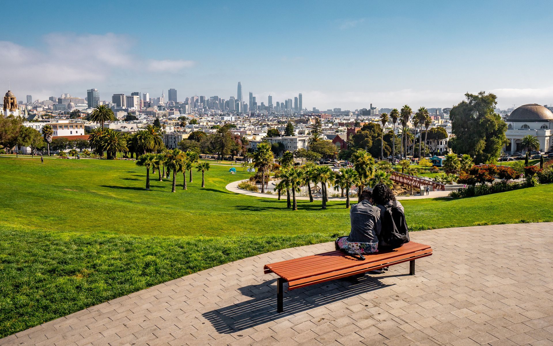 A couple is sitting on a bench in a park looking at the city.