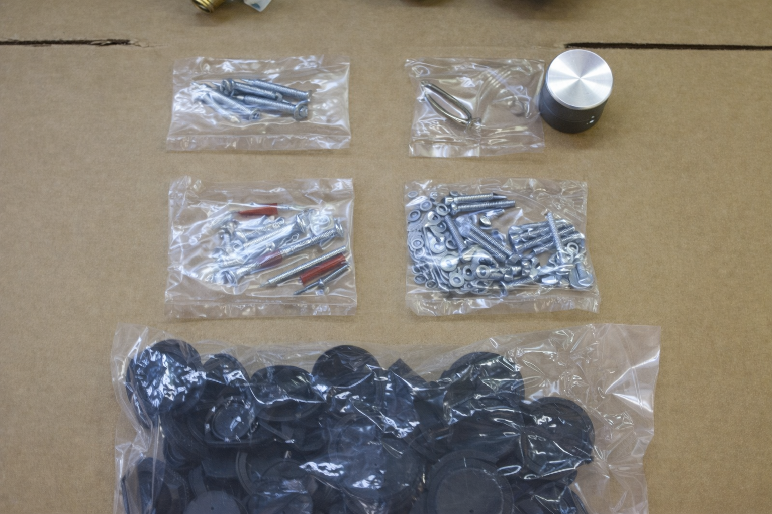 Kit di ferramenta con varie viti, elementi di fissaggio e tasselli di gomma in sacchetti di plastica trasparente, disposti su una superficie di cartone.