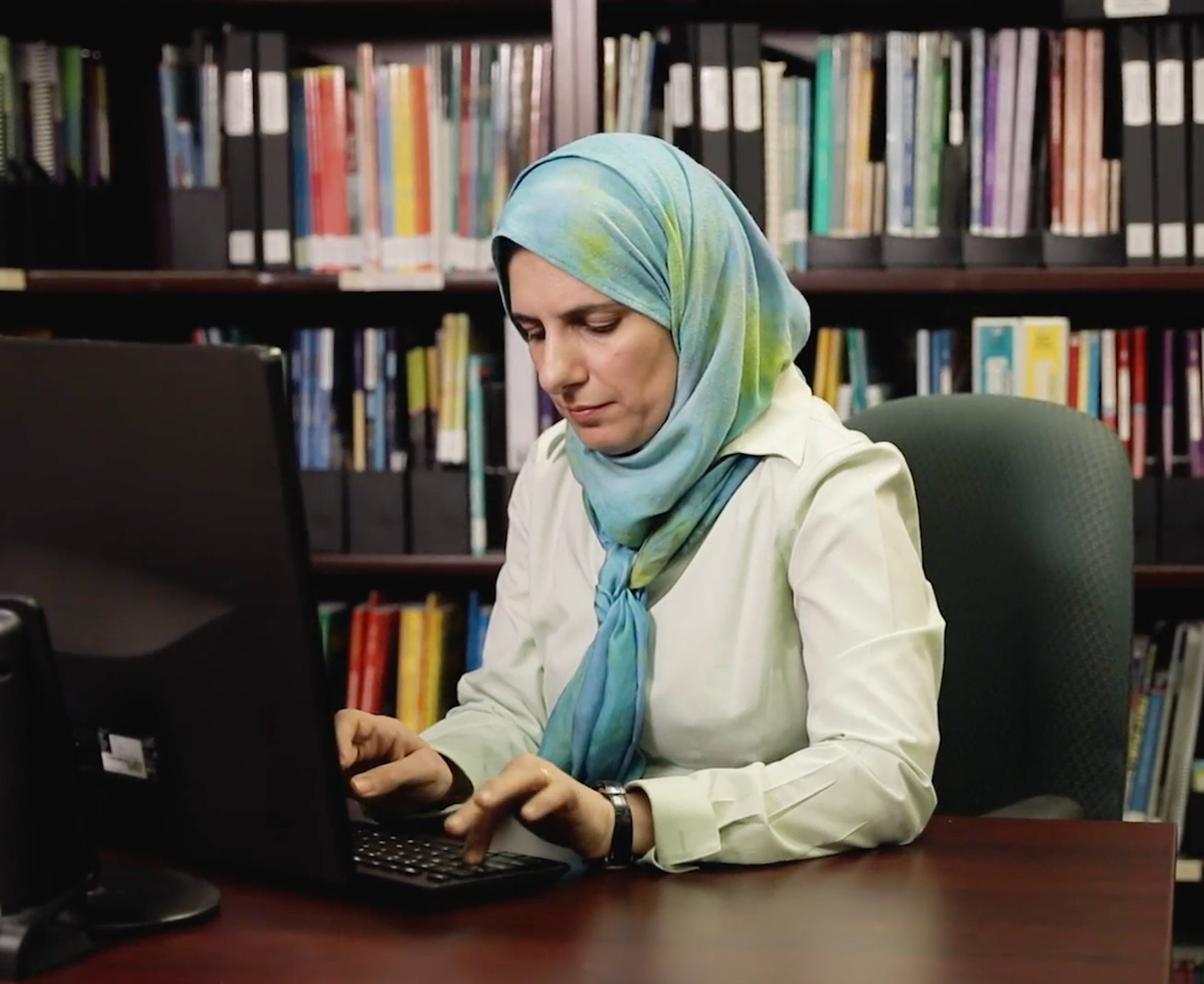 A person wearing a light blue hijab and white shirt sits at a desk in a library, typing on a computer.