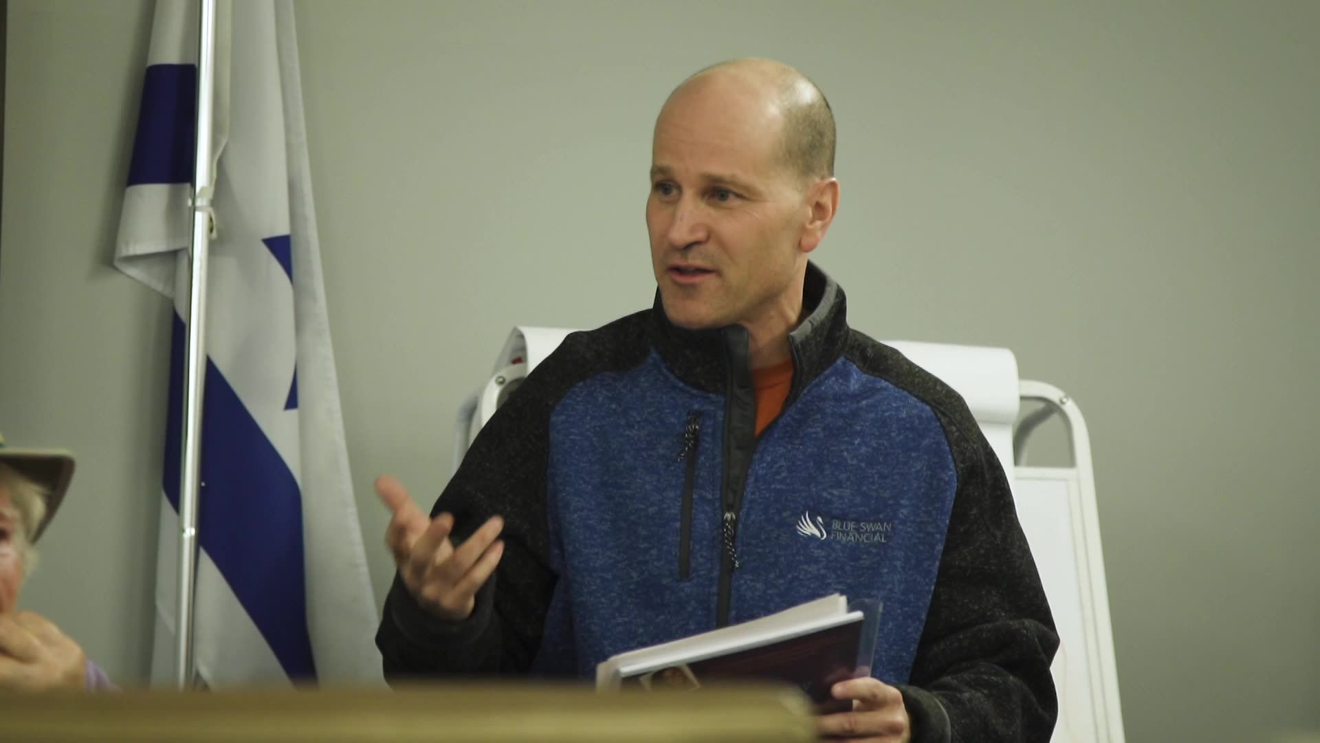 A man in a blue and black pullover gestures while speaking in front of an Israeli flag in an indoor setting.
