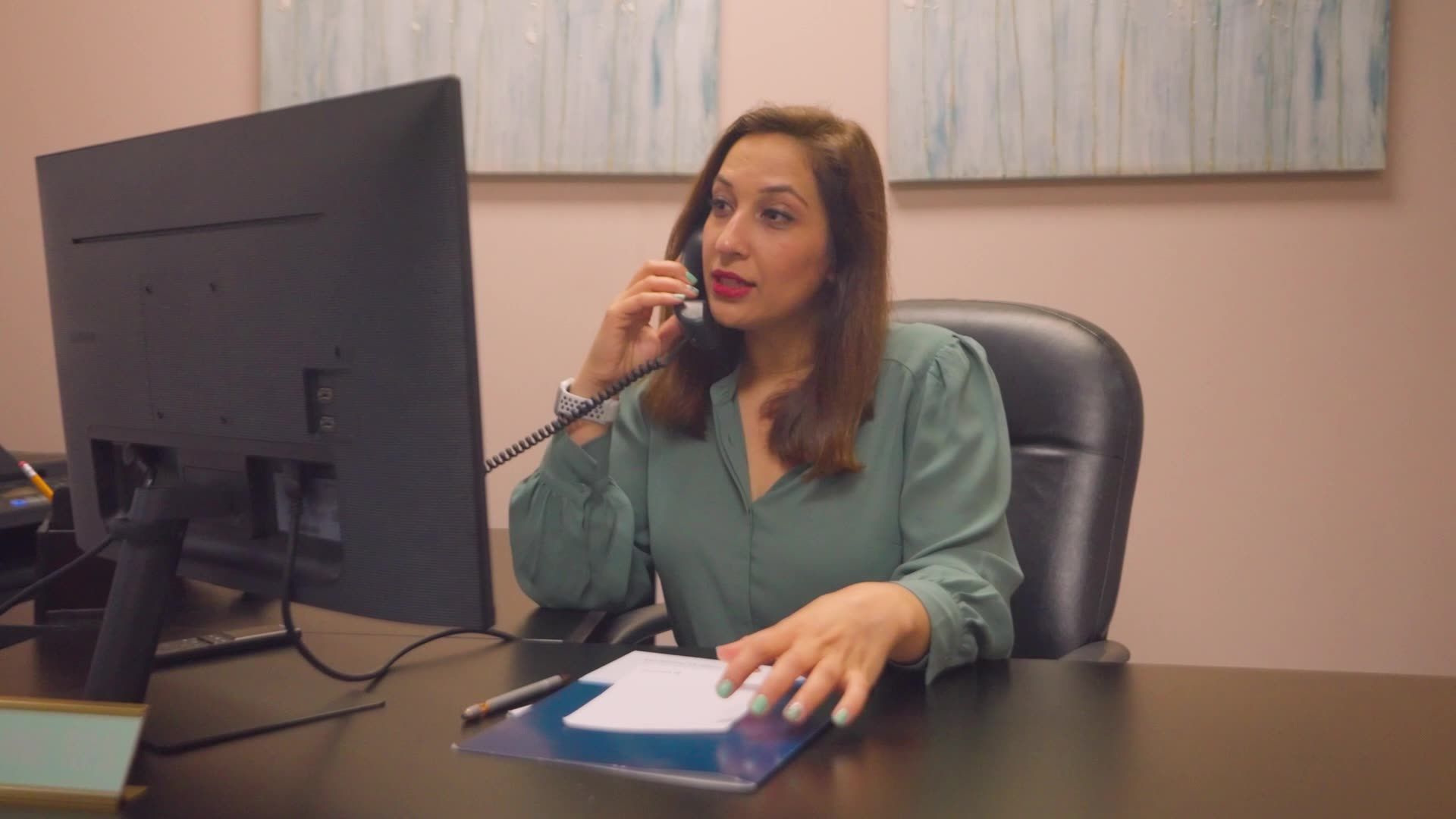 A person in a sage green blouse sits at a desk, looking at a computer screen while talking on a telephone.