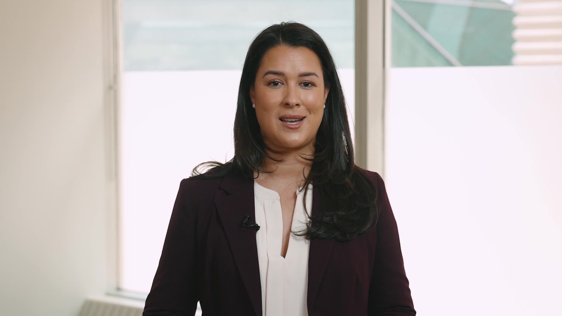 A woman in a pink shirt and black jacket is smiling in front of a window.