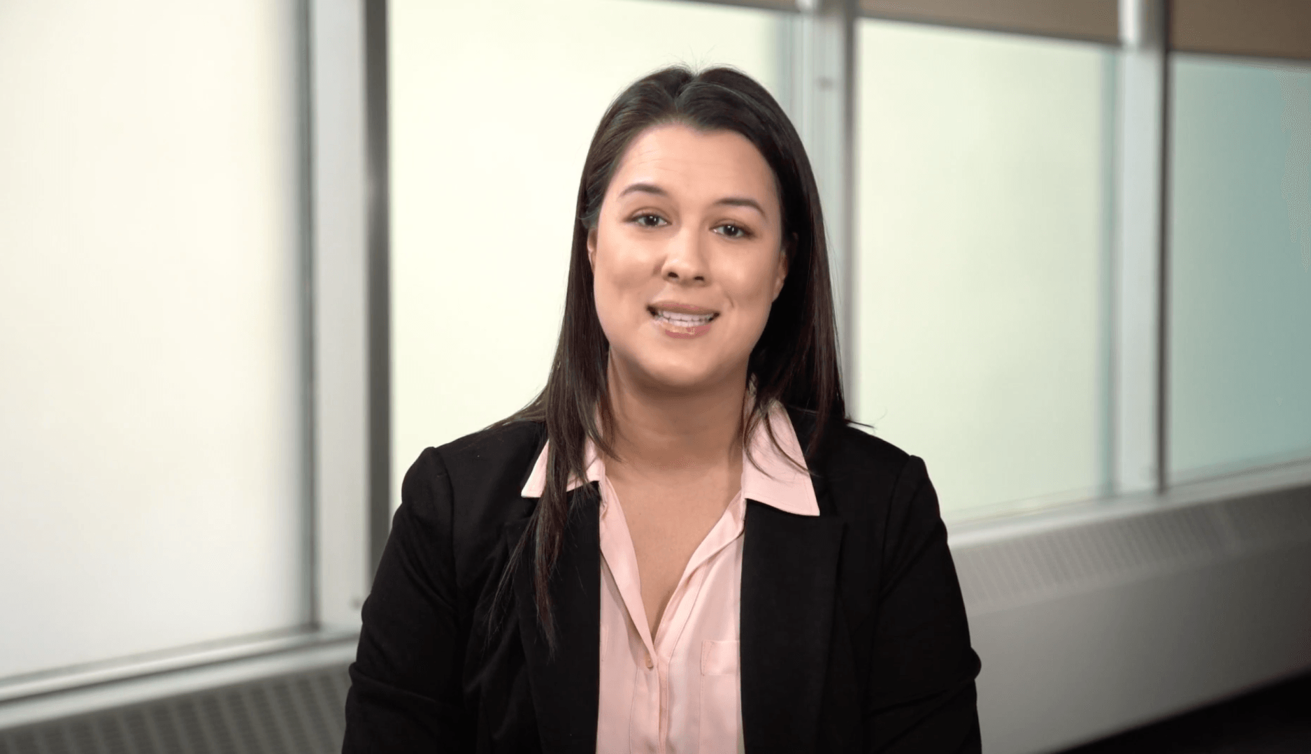A woman in a pink shirt and black jacket is smiling in front of a window.