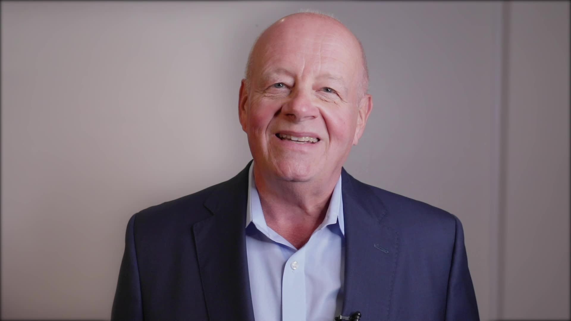 A smiling person in a blue button-down shirt and dark blazer posing against a plain wall.