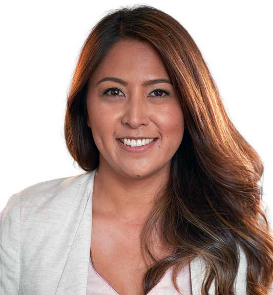 Smiling woman with long brown hair, wearing a light-colored blazer over a pink top, against a plain white background.