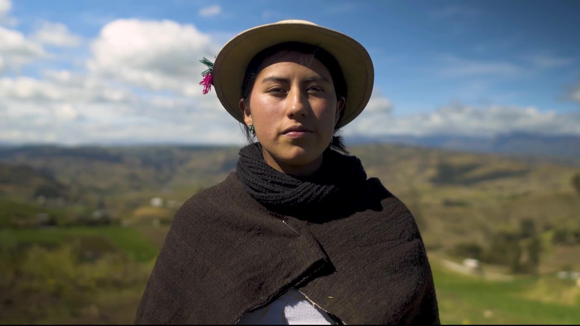 A person wearing a wide-brimmed straw hat and a dark wool shawl stands before a backdrop of hills under a bright sky.