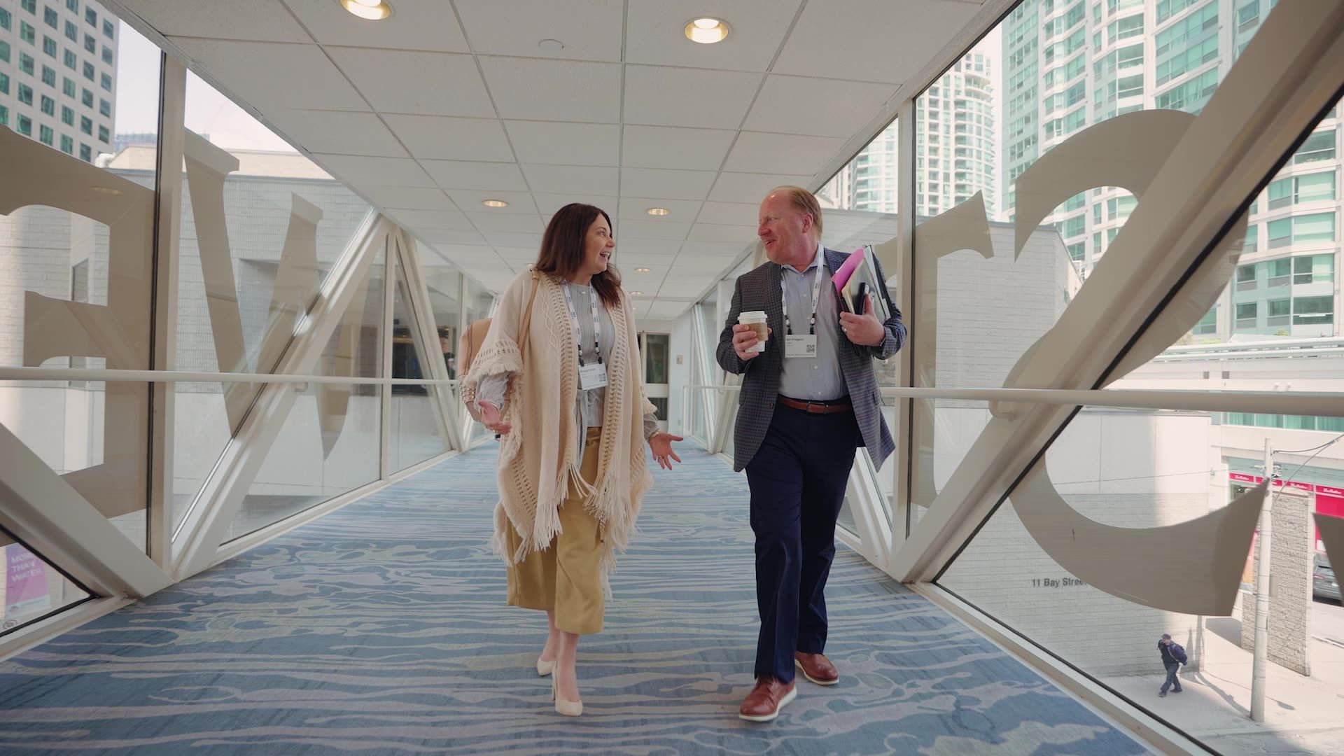 Two people walking and talking in a glass-enclosed pedestrian bridge walkway with large white letters on the windows.