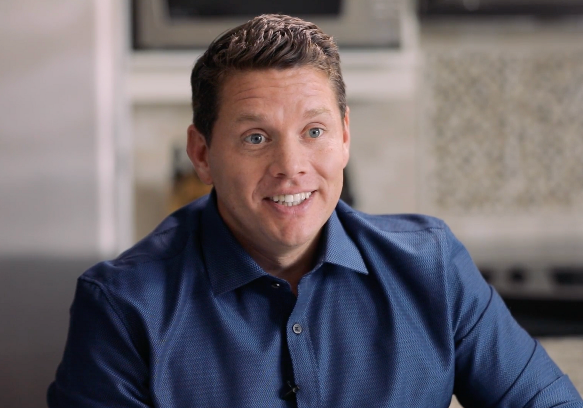 A person with short brown hair in a blue button-down shirt smiling while indoors.