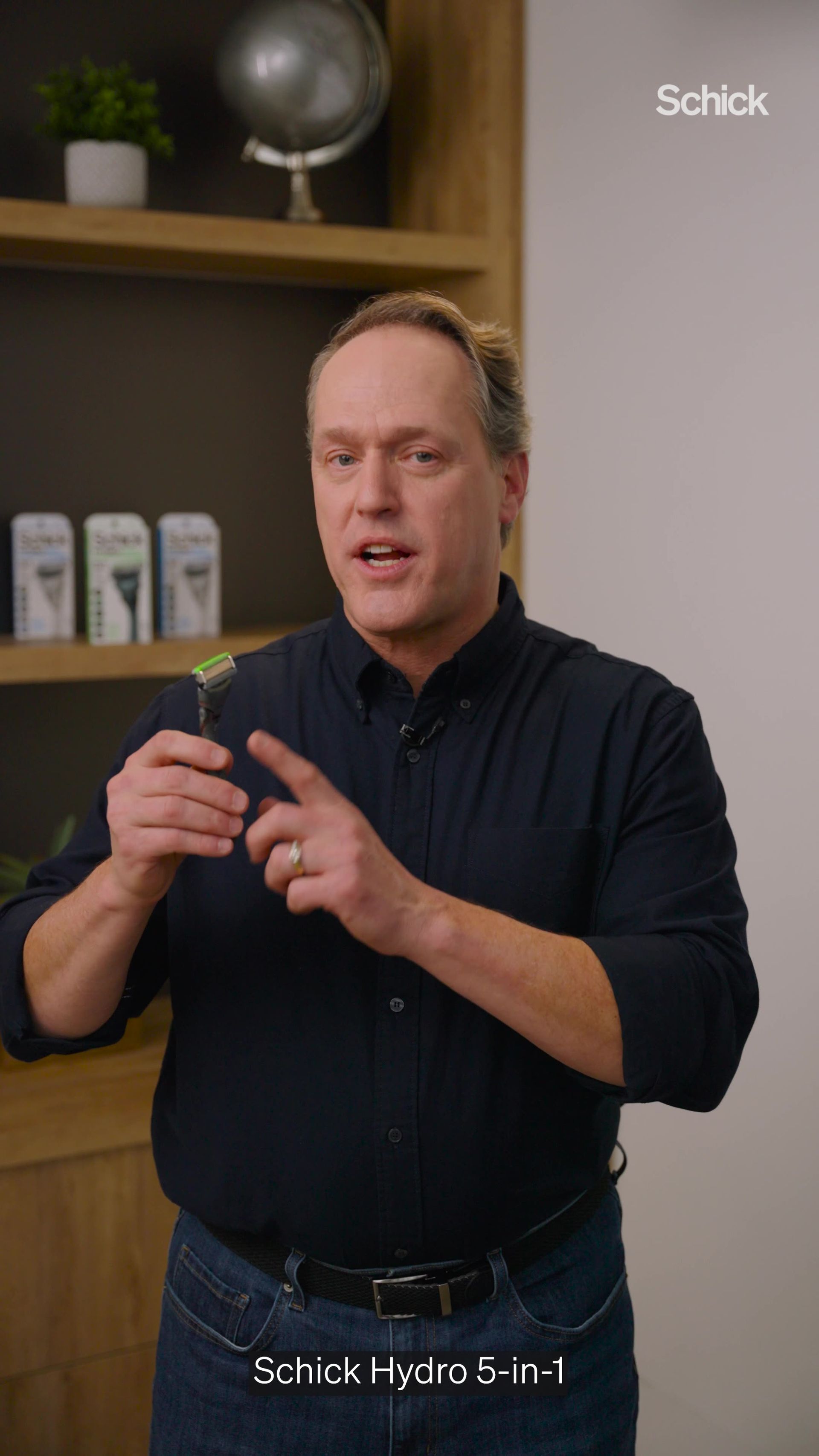 A person in a dark shirt demonstrates a Schick Hydro 5-in-1 razor in front of a shelf.