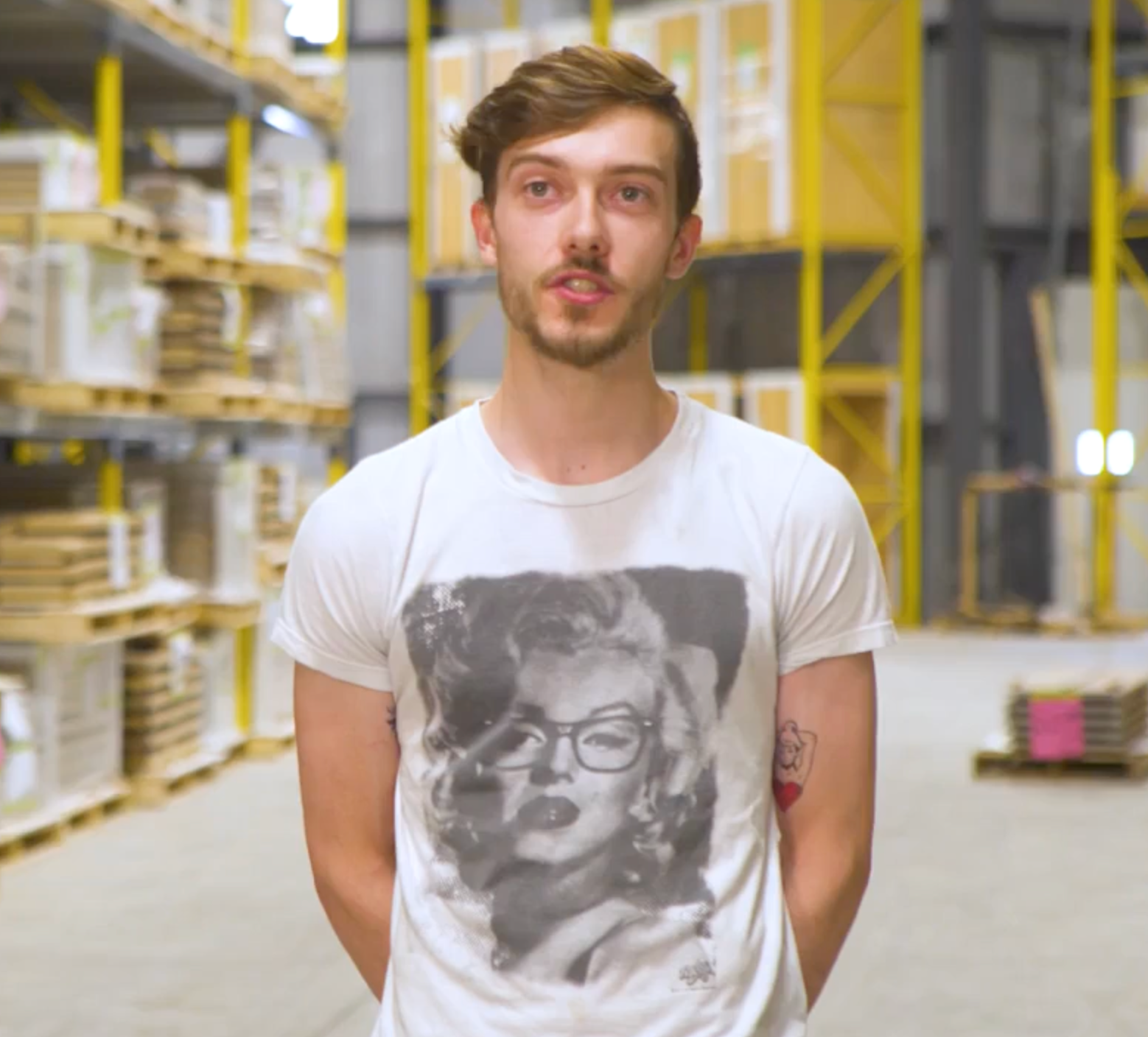 A person wearing a white Marilyn Monroe graphic t-shirt stands in a brightly lit industrial warehouse with pallet racking.