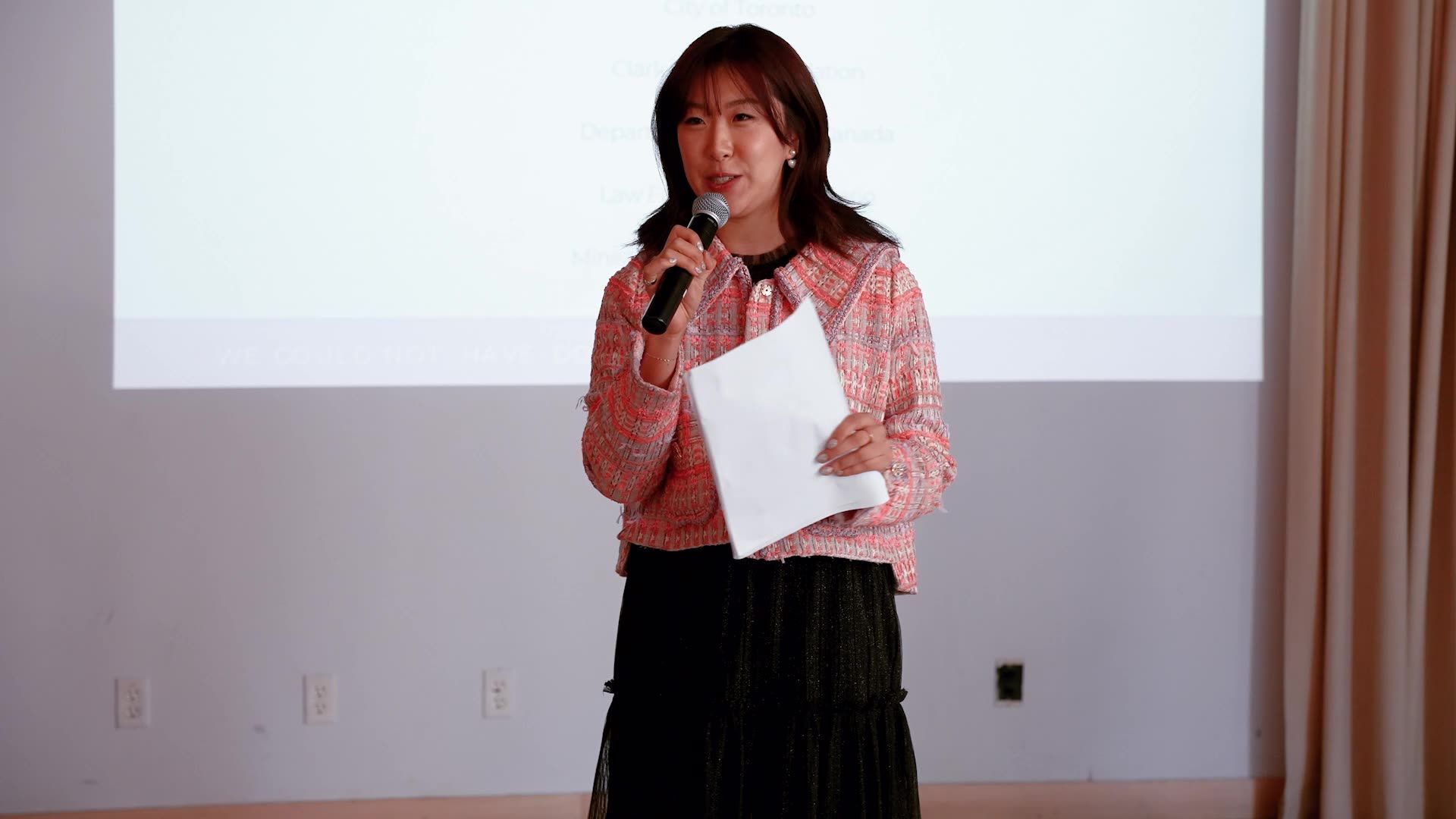 A speaker in a pink tweed jacket stands on stage holding a microphone and papers in front of a white projection screen.
