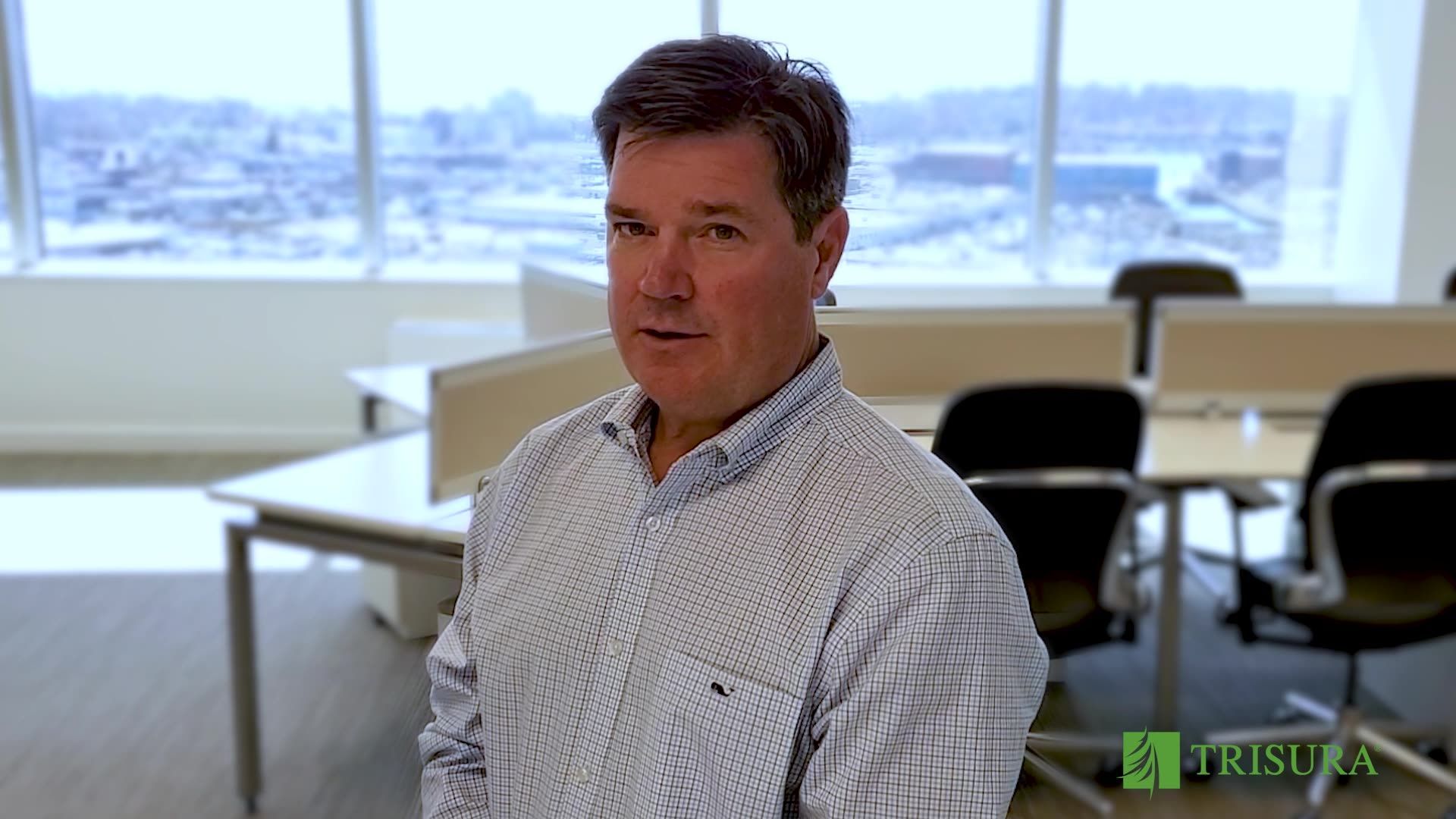 A person in a light-patterned shirt stands in a brightly lit, modern office with desks and windows in the background.