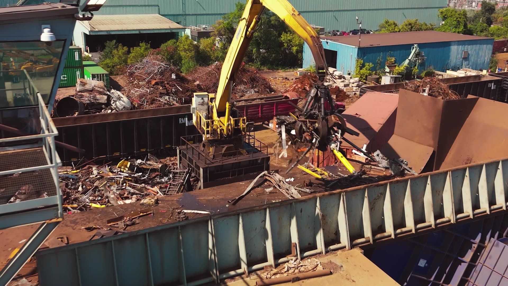 A yellow crane with a mechanical grapple moves scrap metal inside a large industrial container at a recycling yard.