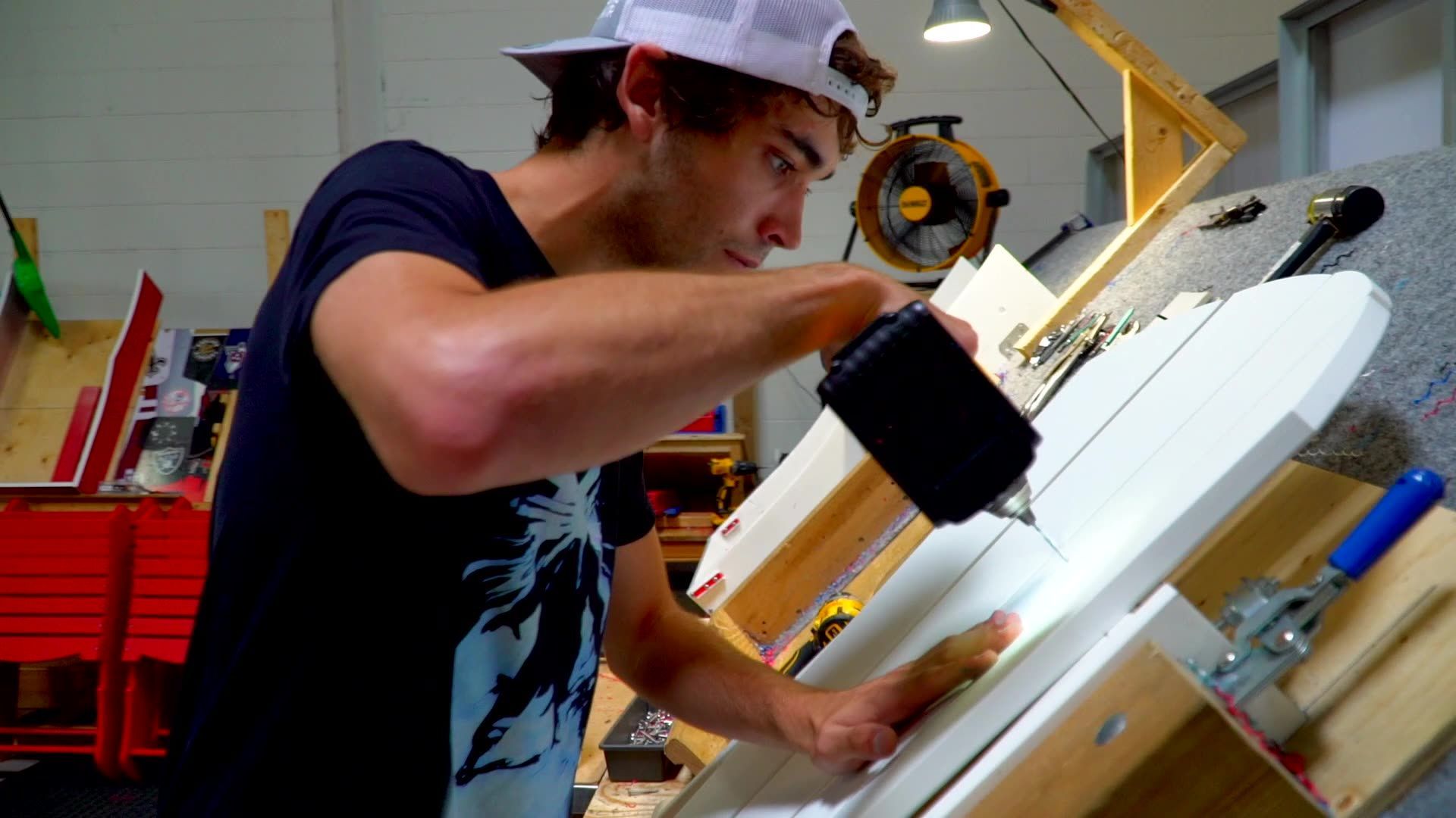 A person in a baseball cap uses a power drill on a white wooden panel mounted on a workbench in a workshop.