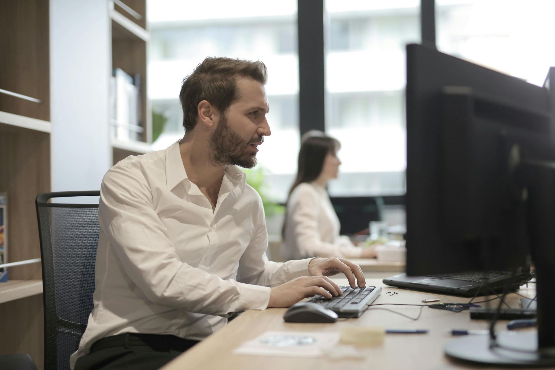 A professional in a white shirt types on a keyboard while working at a desk in a bright, modern office.