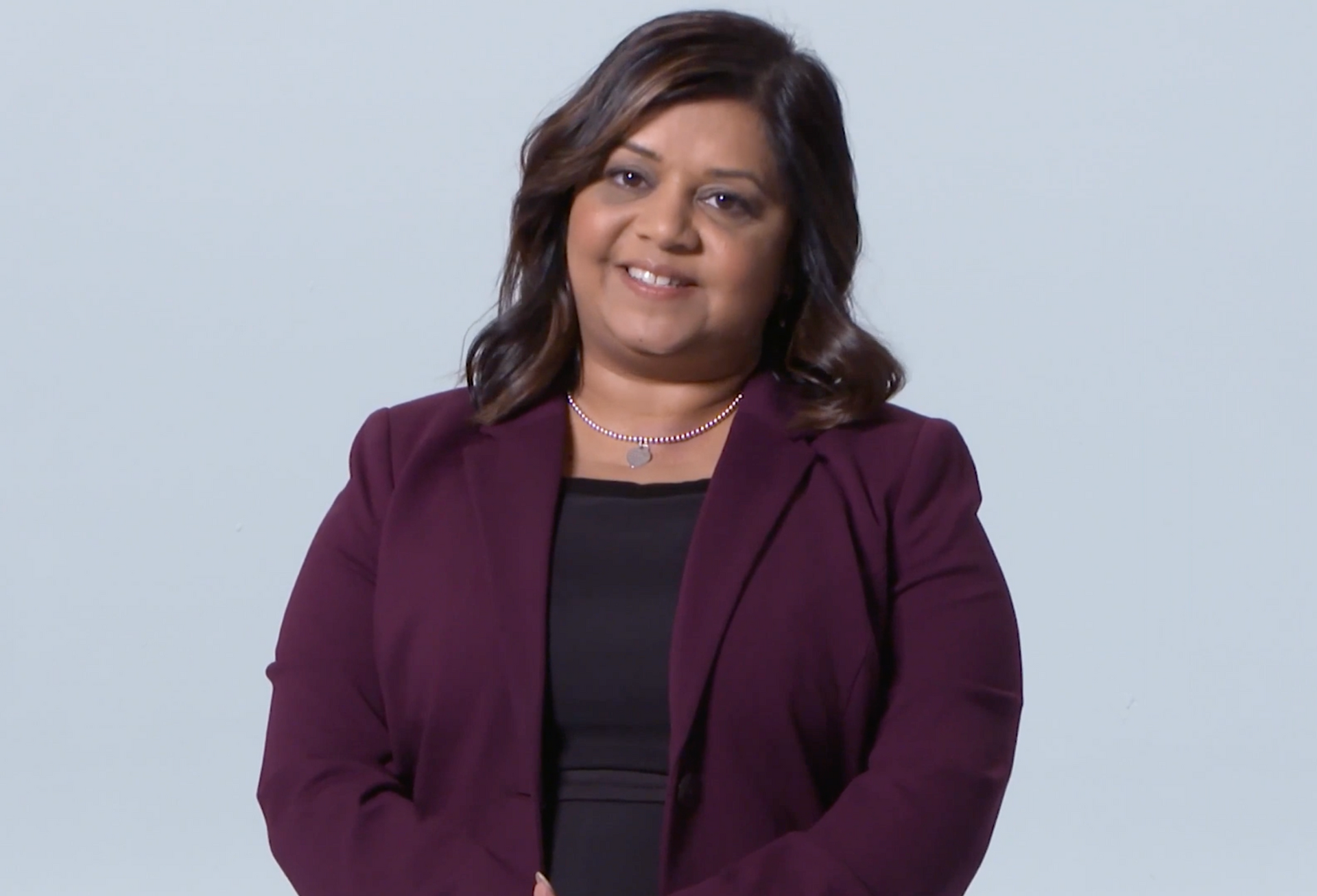 A person with dark hair wears a plum-colored blazer over a black top, smiling against a light gray background.
