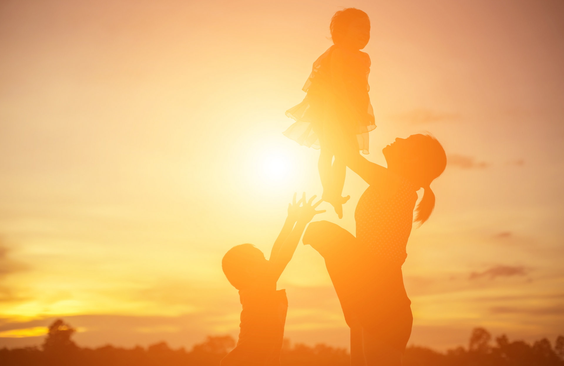 Silhouetted family playing: Mother throwing child in the air as another child watches against a sunset.