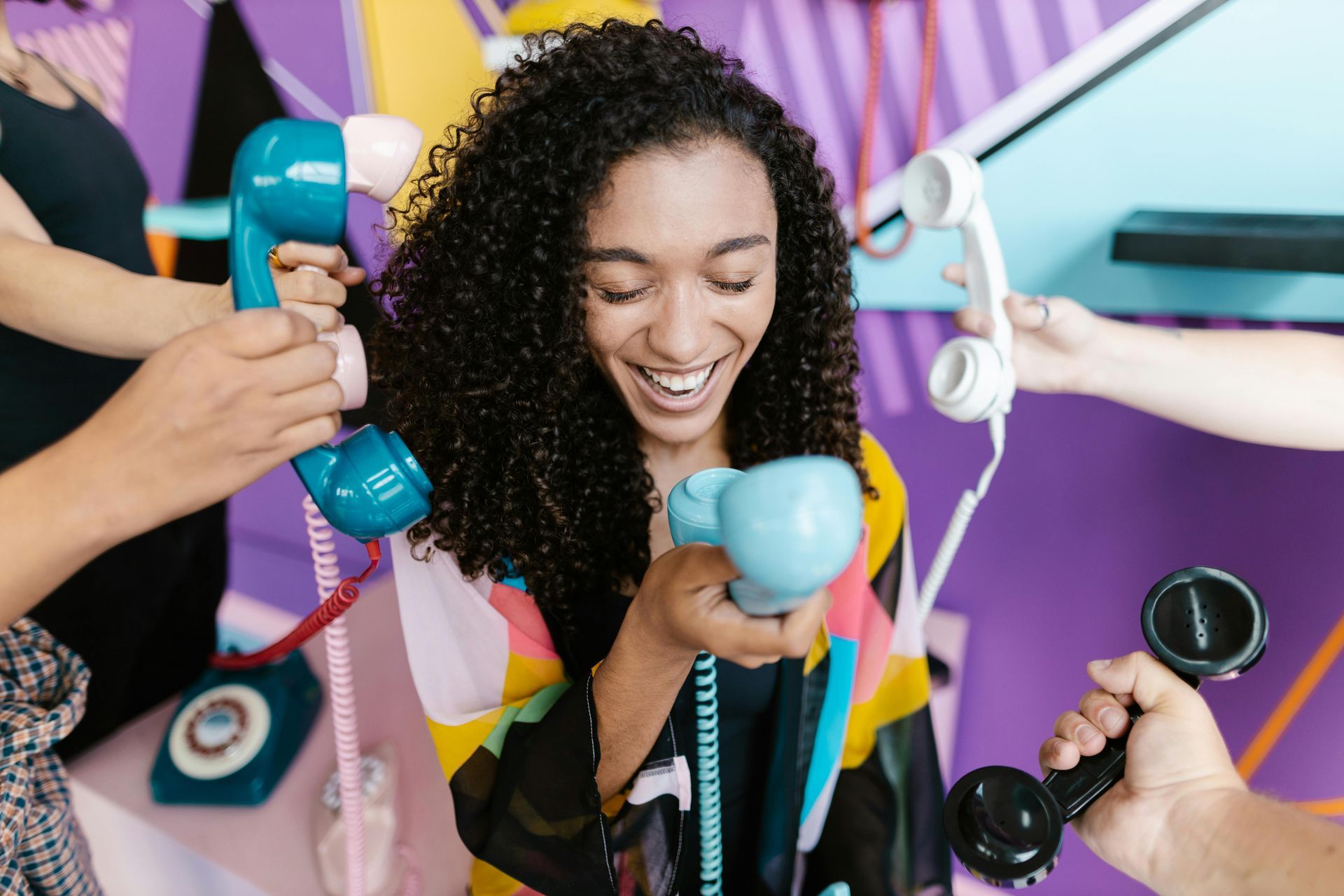 A person laughing as multiple colorful vintage telephone handsets are held up to them from various sides.