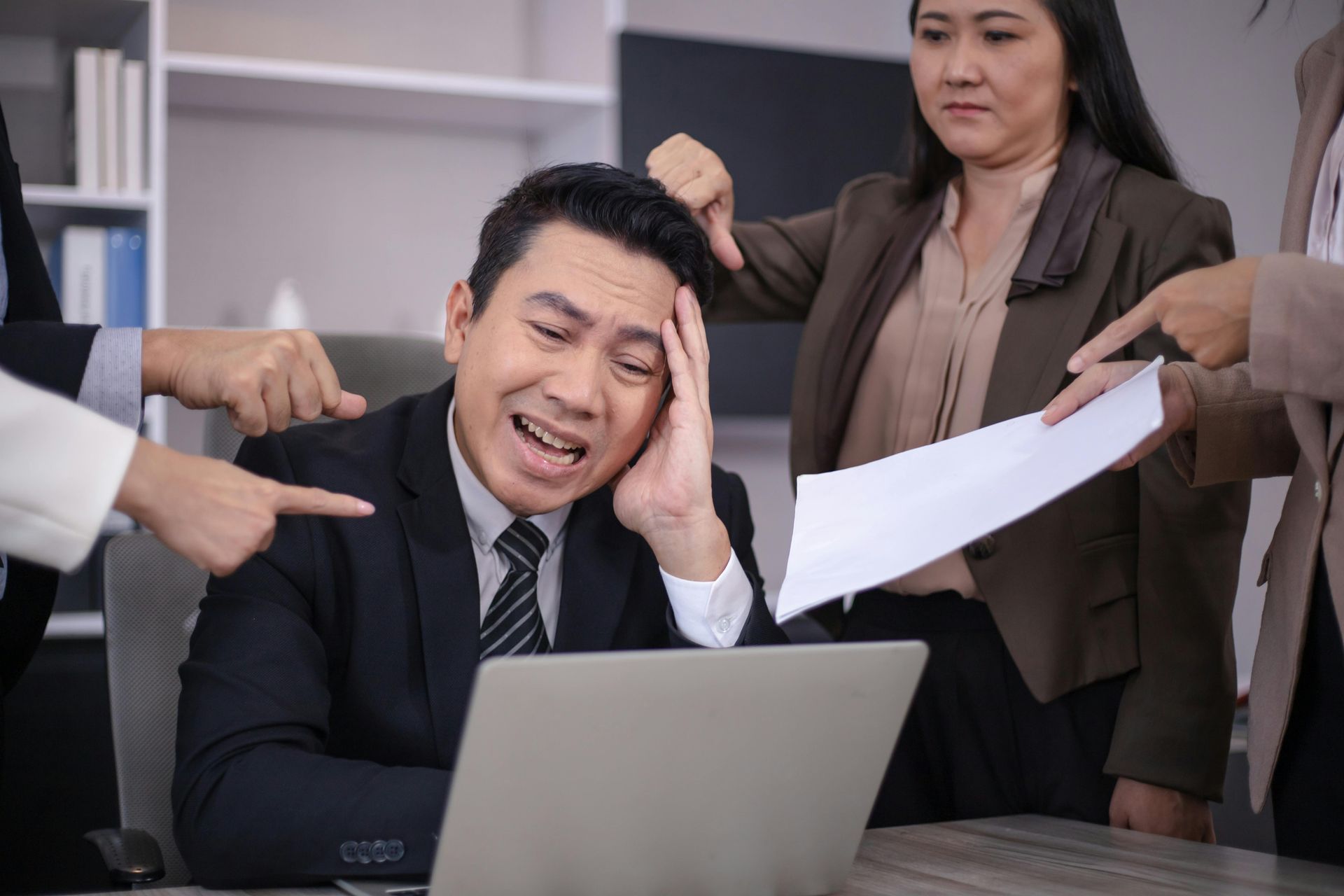 Man in suit, stressed at desk, being pointed at by colleagues in office.