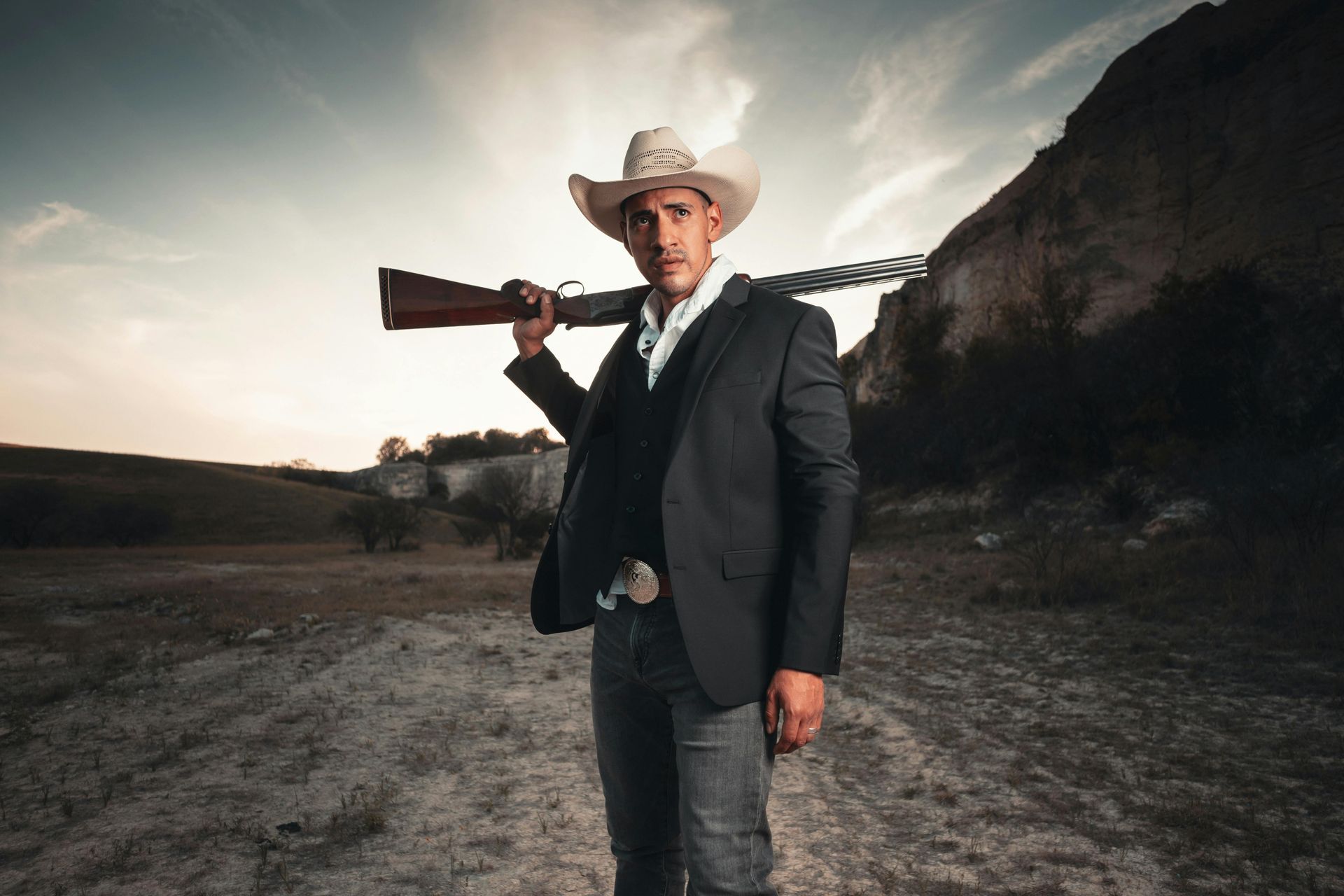 Cowboy in a suit and hat carrying a rifle, standing outdoors against a rocky backdrop.