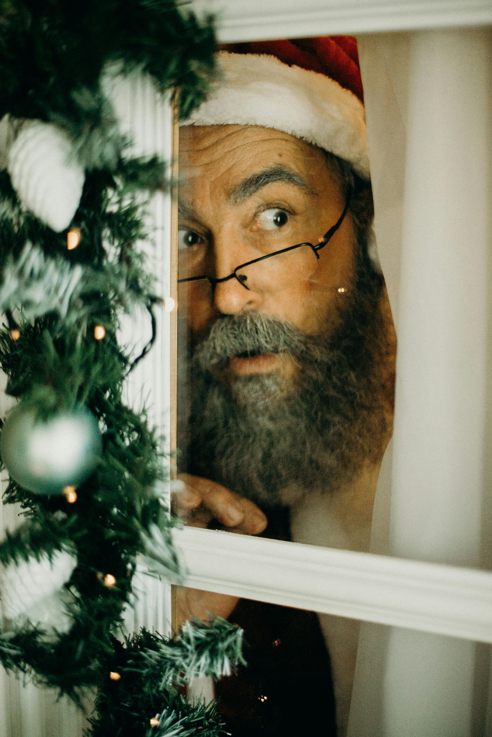 Santa Claus peers through a window, surprised, decorated with a Christmas garland.