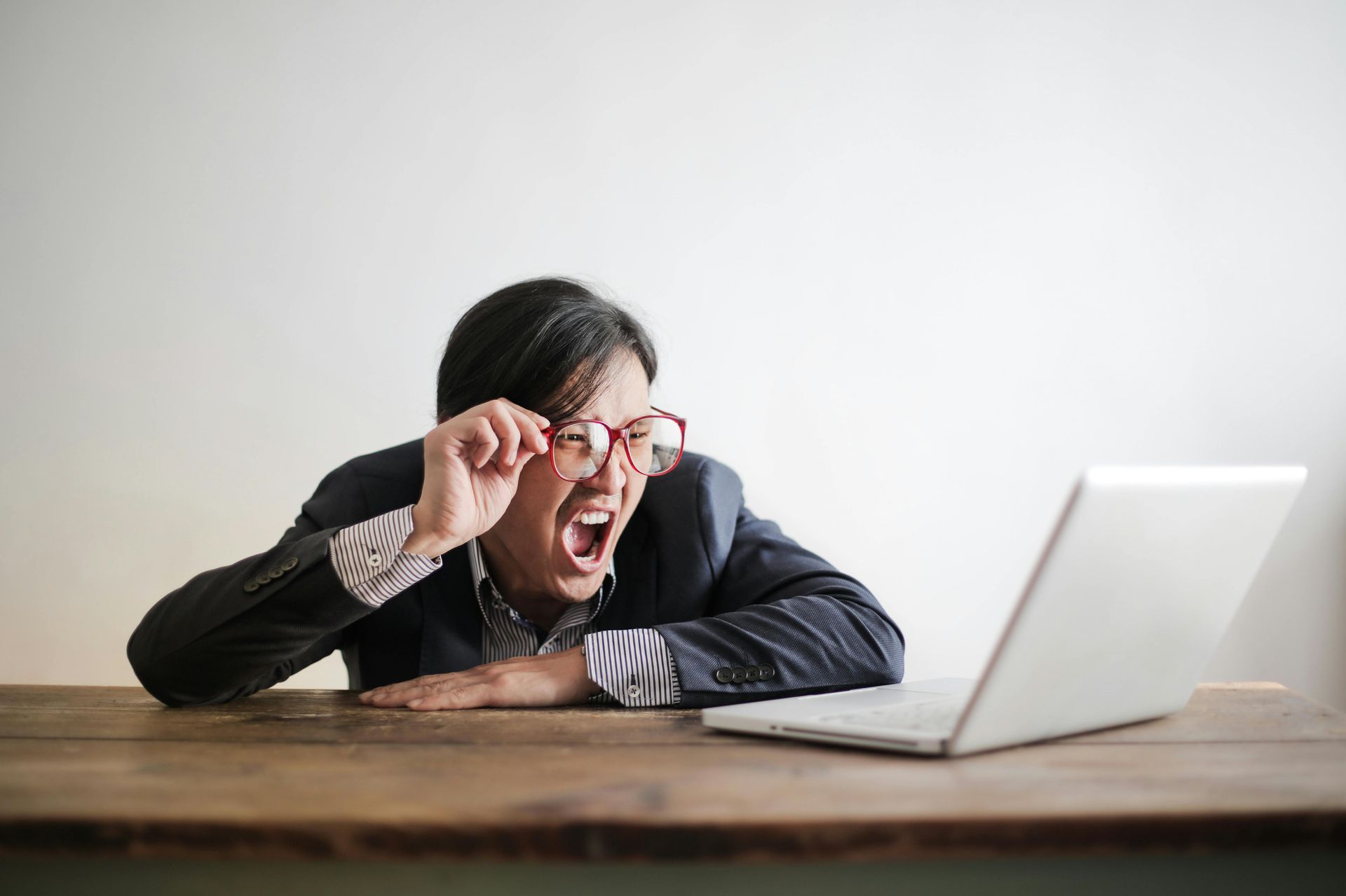 Man in blazer leans toward laptop, eyes wide, mouth open, holding glasses; table and white wall in background.