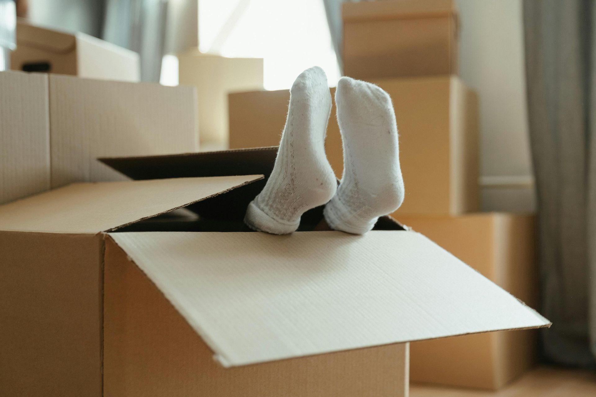 Feet in white socks sticking out of a cardboard box, surrounded by other packed boxes.