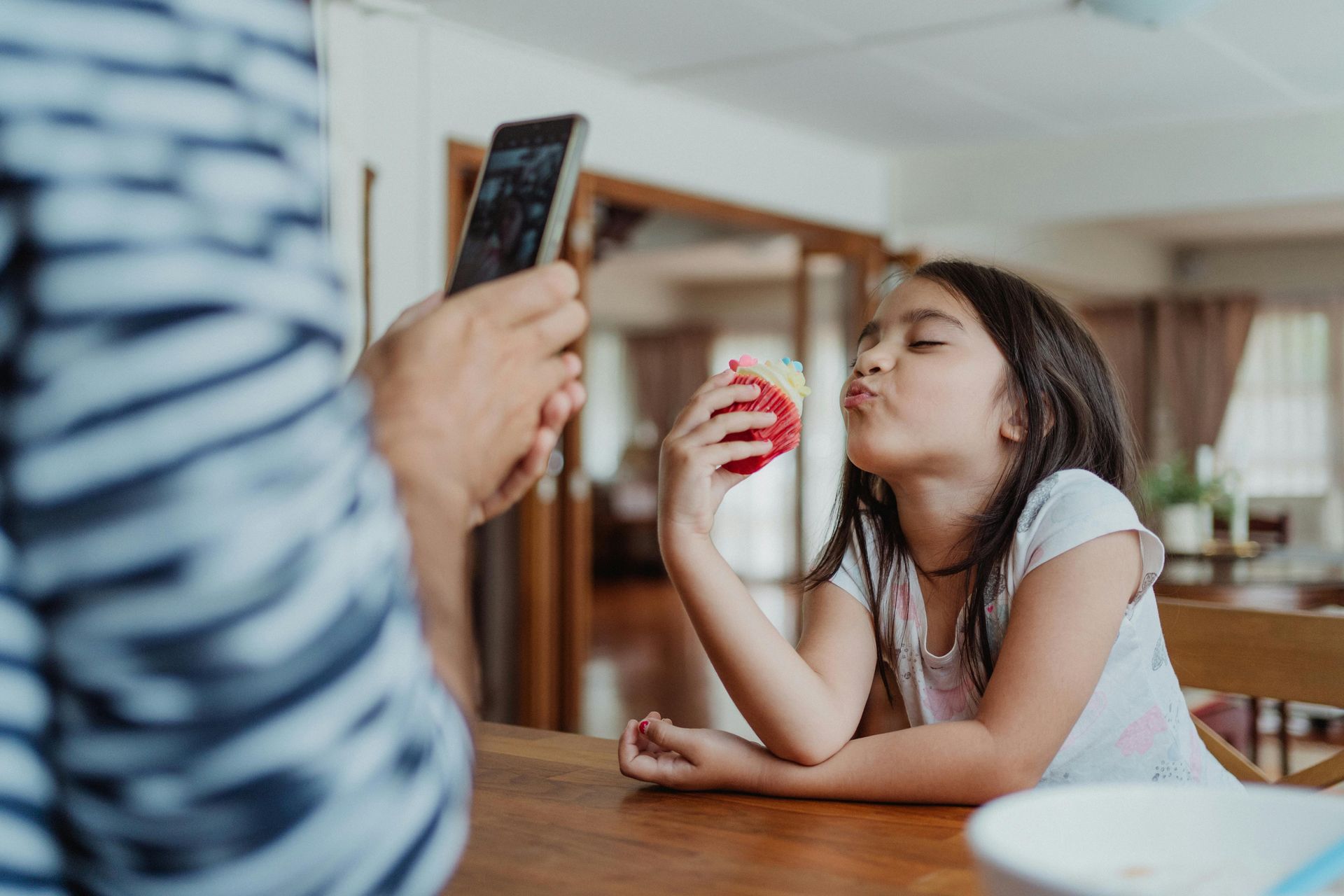 A little girl is sitting at a table blowing bubbles while a man takes a picture of her.