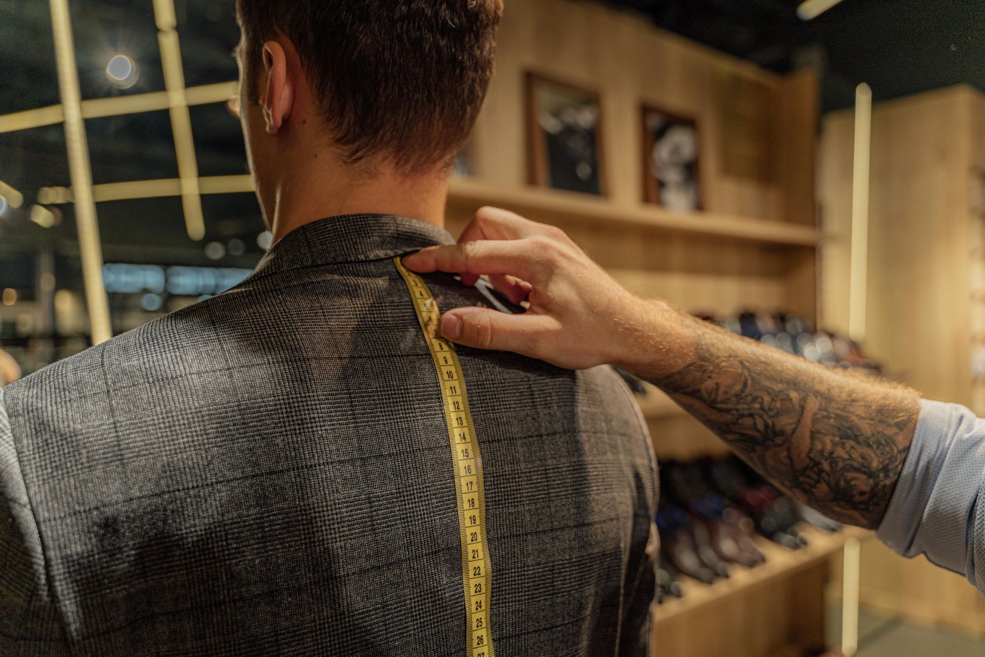 Tailor measuring the back length of a man's jacket with a yellow tape measure in a shop.