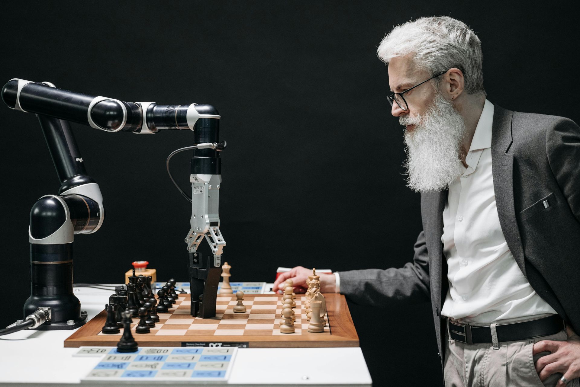 A person with a beard and suit plays a game of chess against a robotic arm.