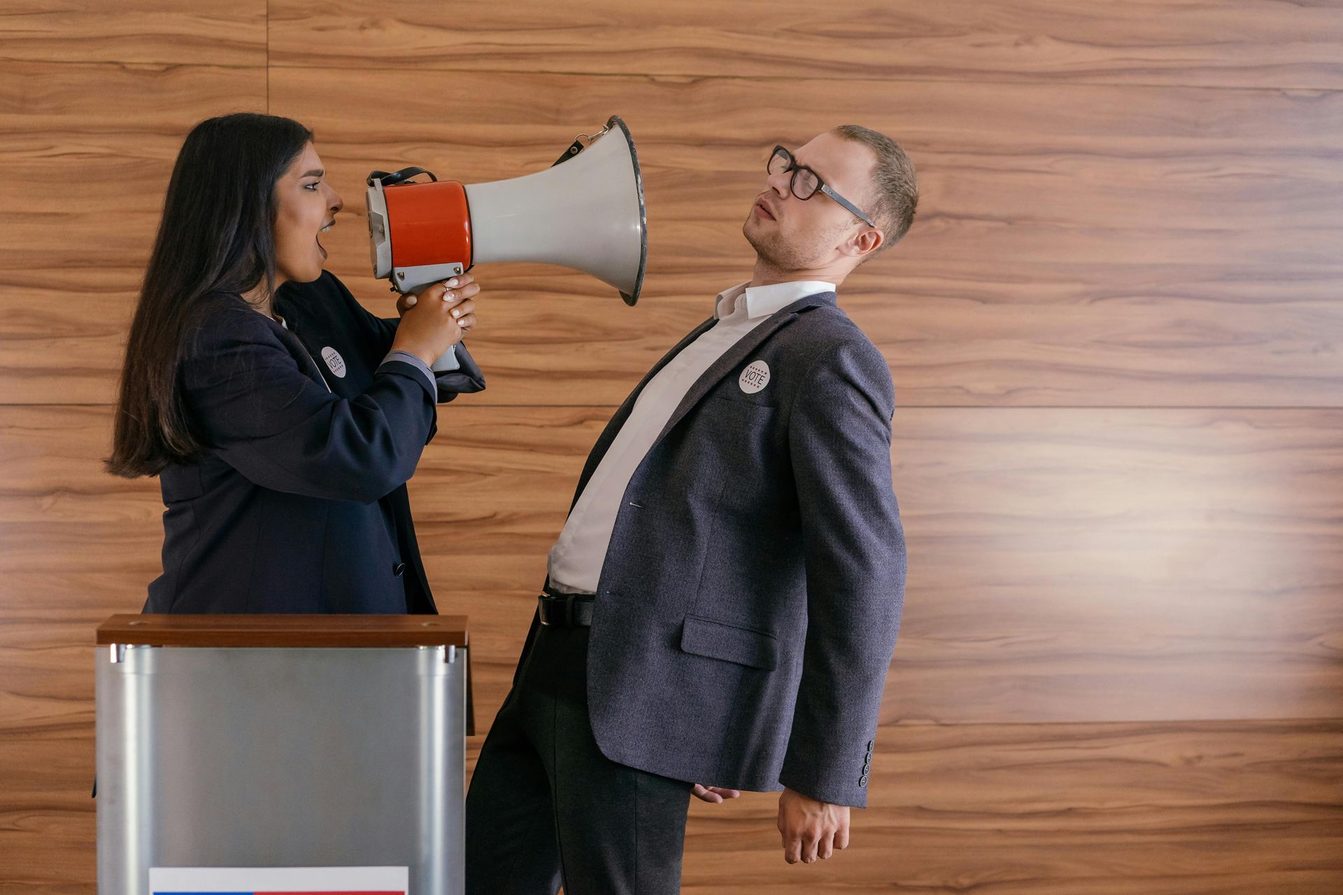 Woman yelling into megaphone at man; both wear suits, stand near podium, wood background.