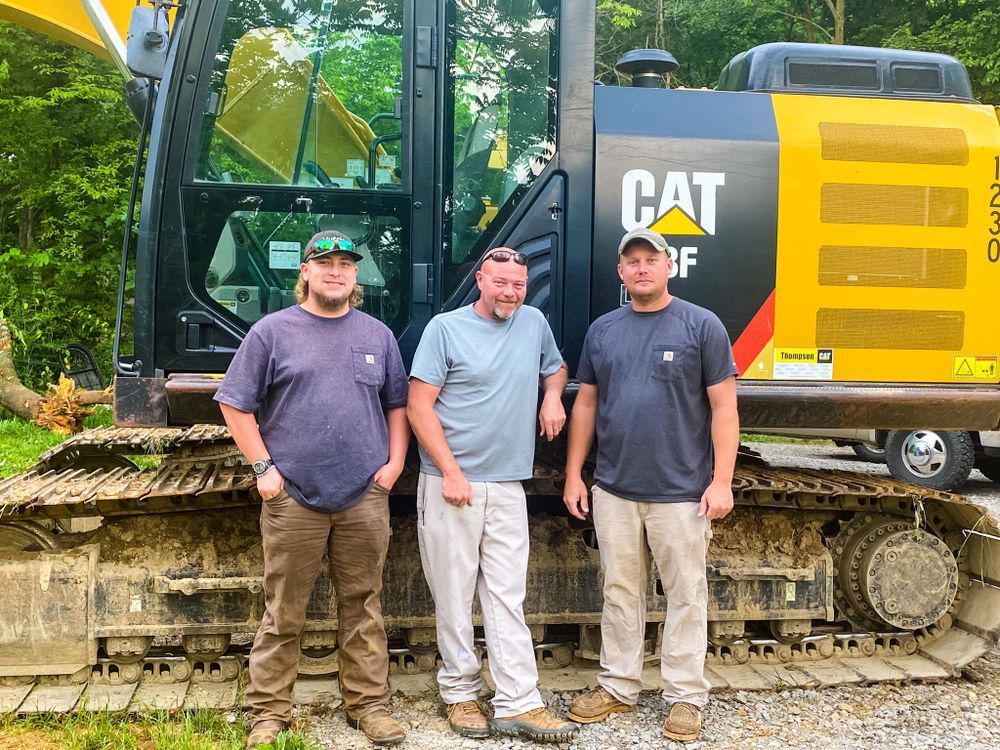 Three men in casual work clothes stand smiling in front of a yellow Caterpillar excavator parked on a gravel lot.