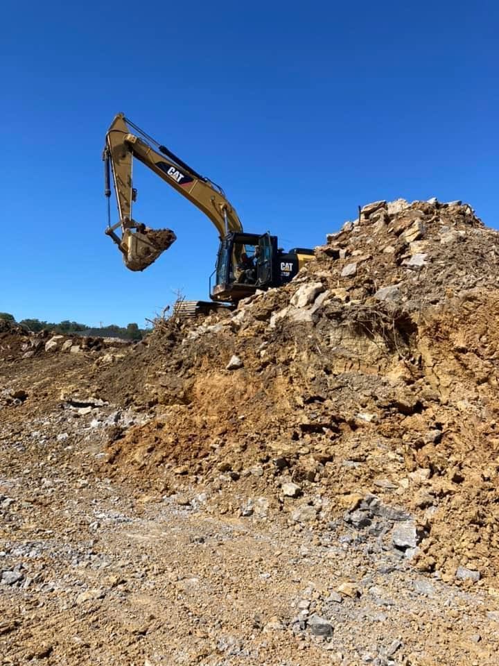A yellow CAT excavator sits on top of a rocky dirt mound against a clear blue sky.