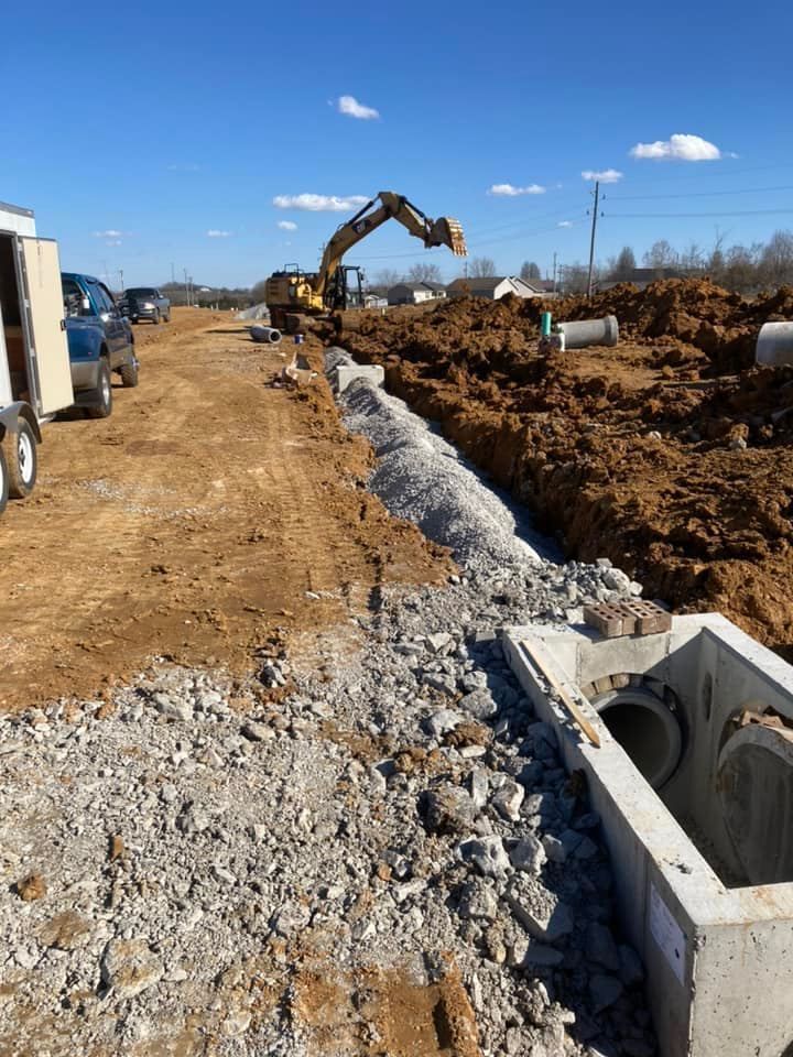 An excavator works on a construction site with concrete drainage pipes and a trench under a clear blue sky.