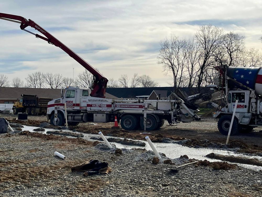 A concrete pump truck and a cement mixer truck at a construction site with gravel and exposed pipes on a cloudy day.