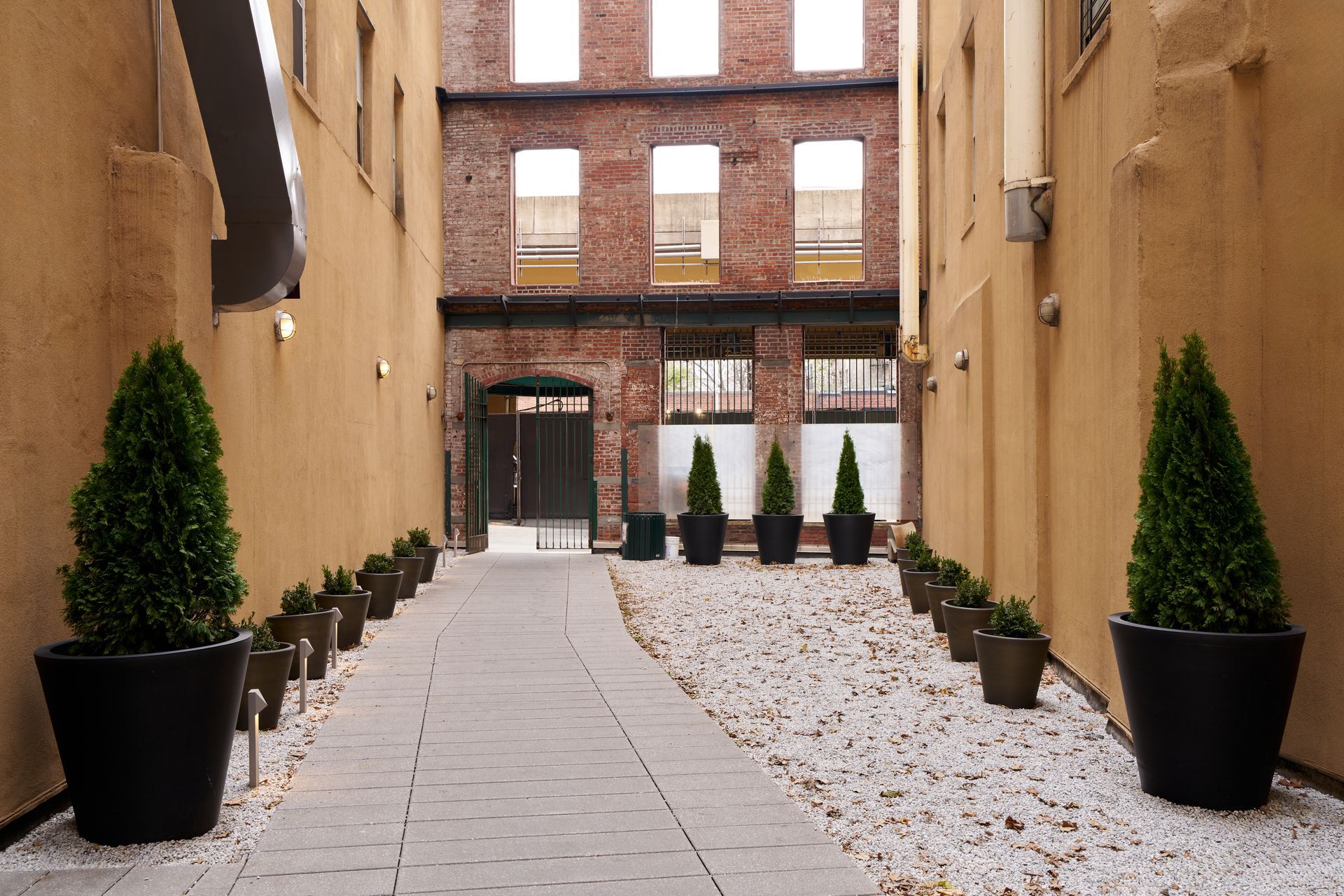 Narrow alleyway with brick building and potted trees. White gravel pathway, beige walls.
