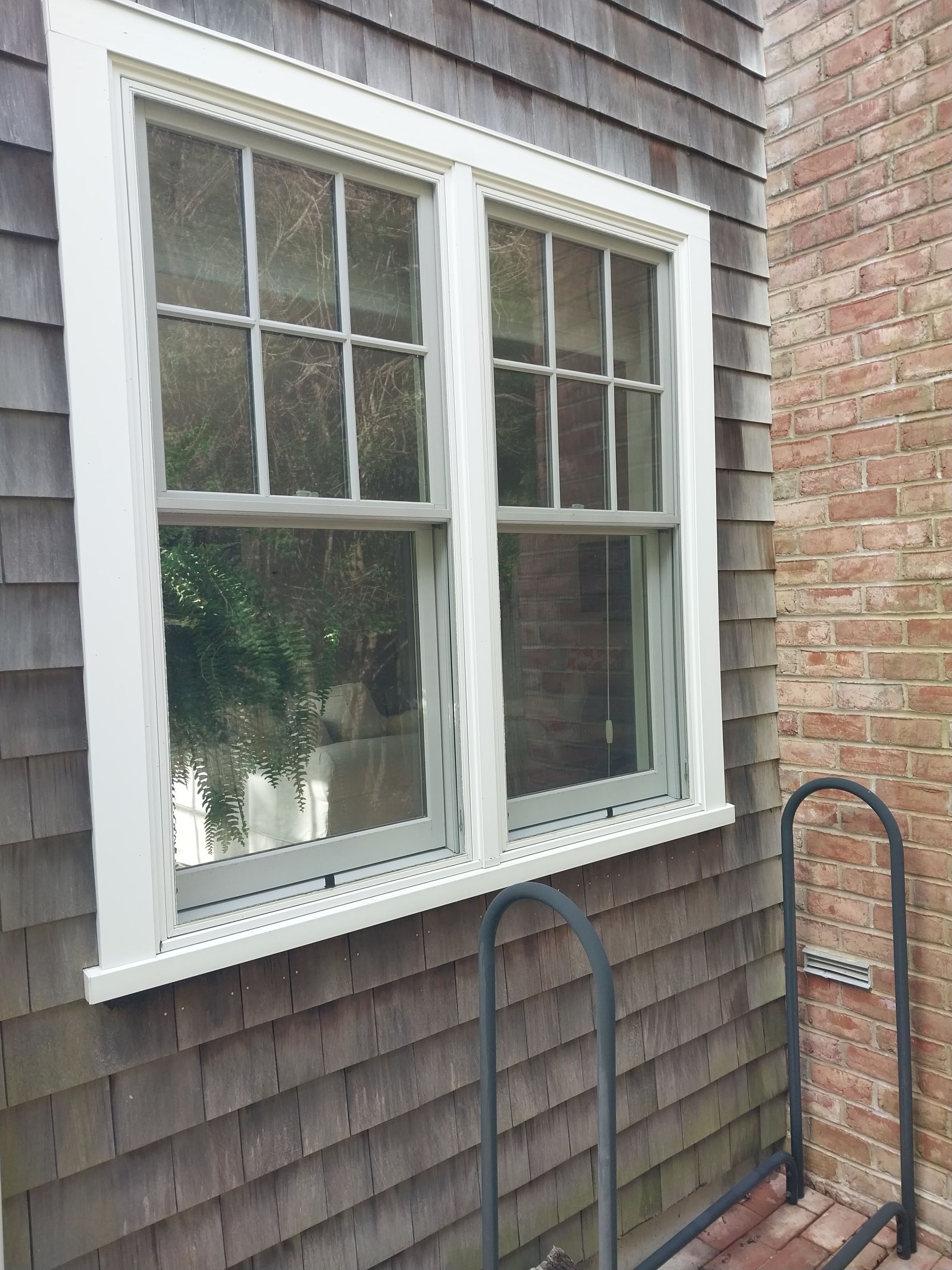 White-framed double-hung windows set into a gray-brown shingled exterior wall, next to a brick wall and a black handrail.