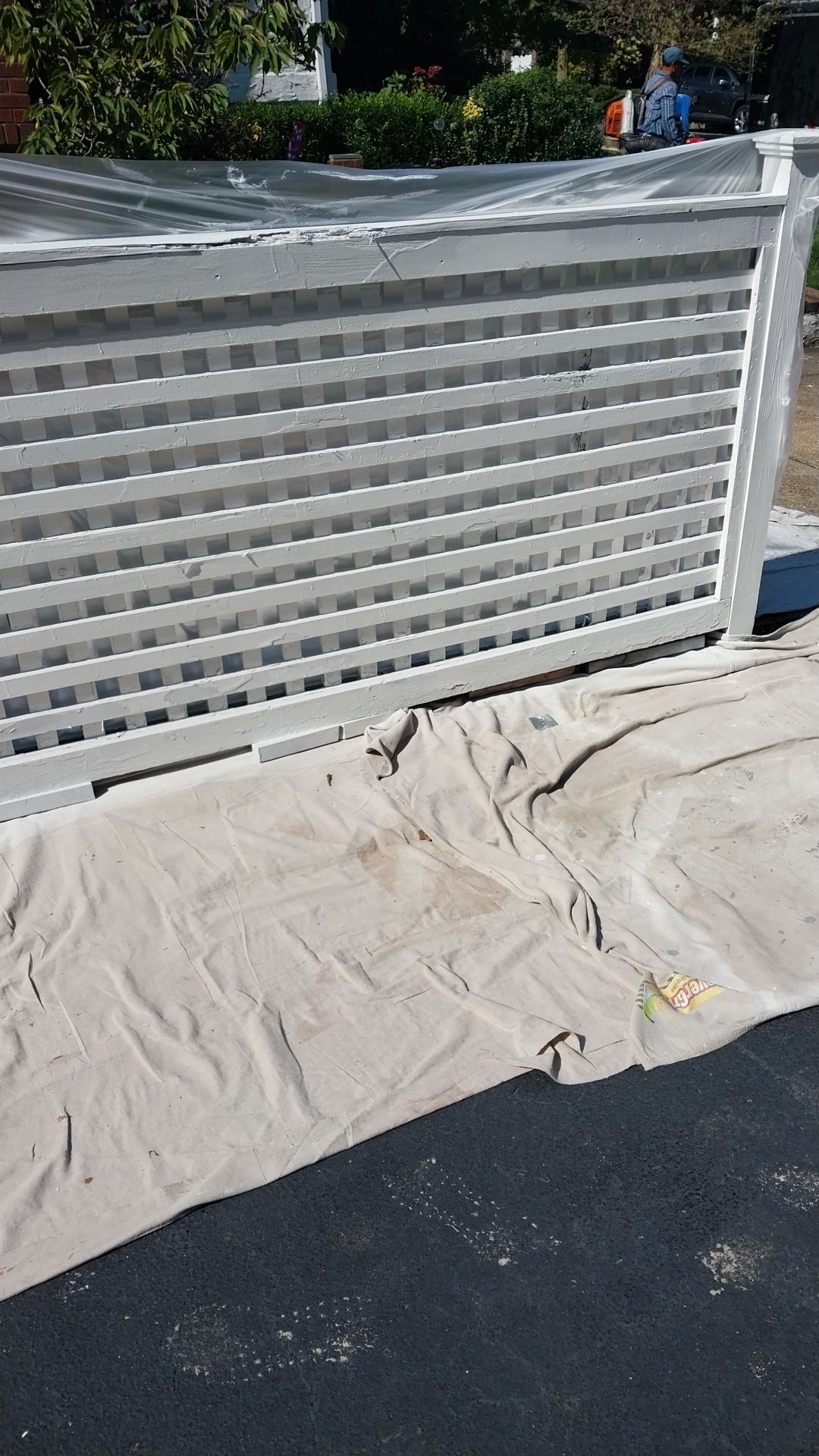 White, horizontal slat fence, partially painted, with a drop cloth on the asphalt below.