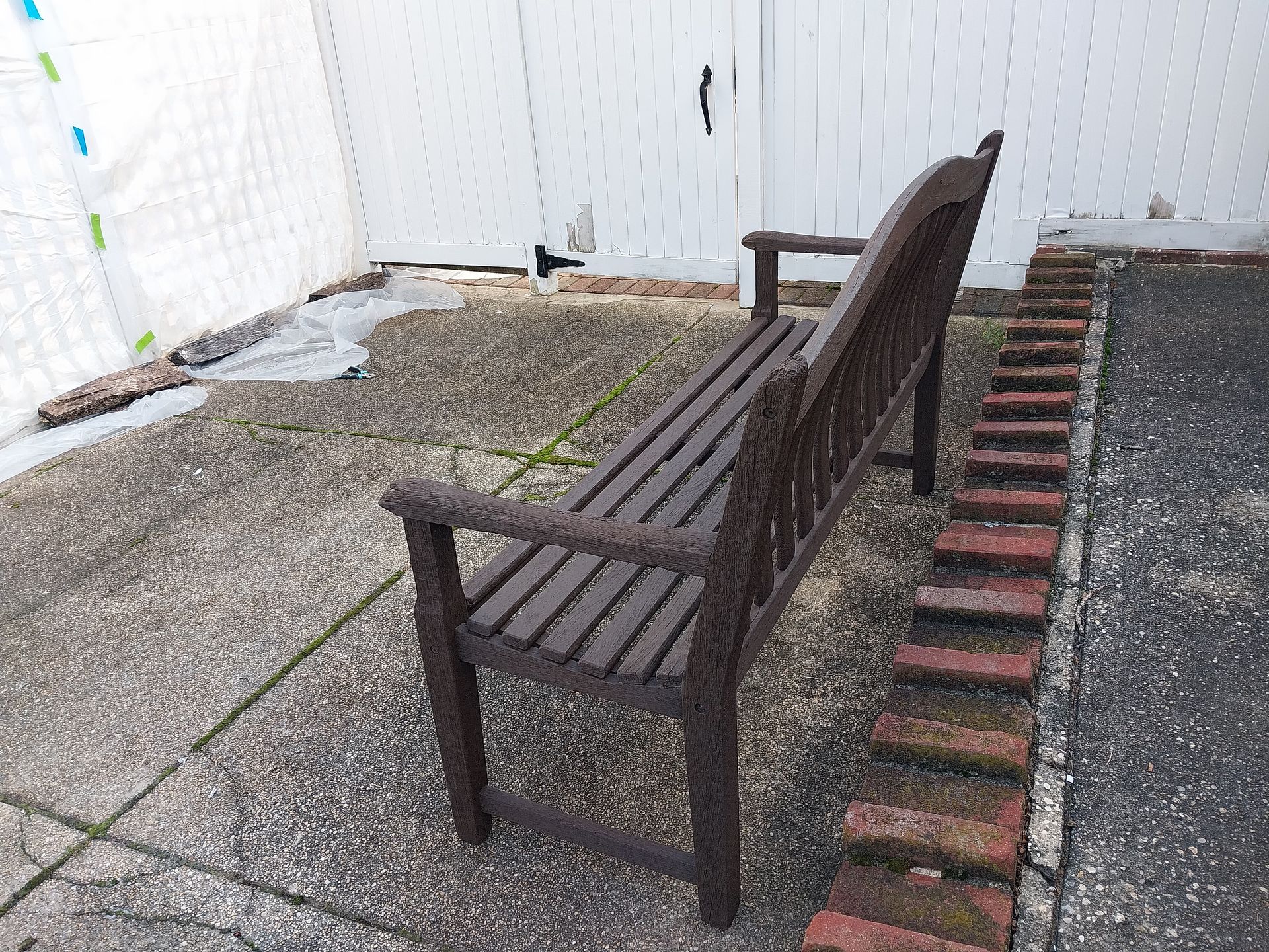 Wooden bench on a concrete patio, positioned near a brick border and white fence.