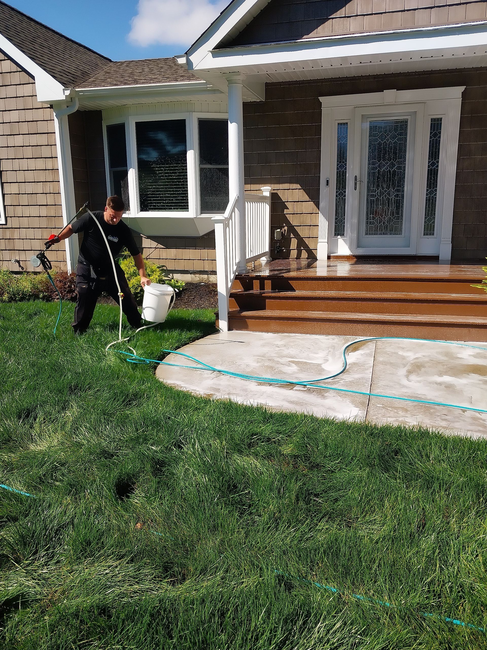 Man washing concrete steps with a hose and bucket; house with bay window and front door in the background, lush green grass in the foreground.